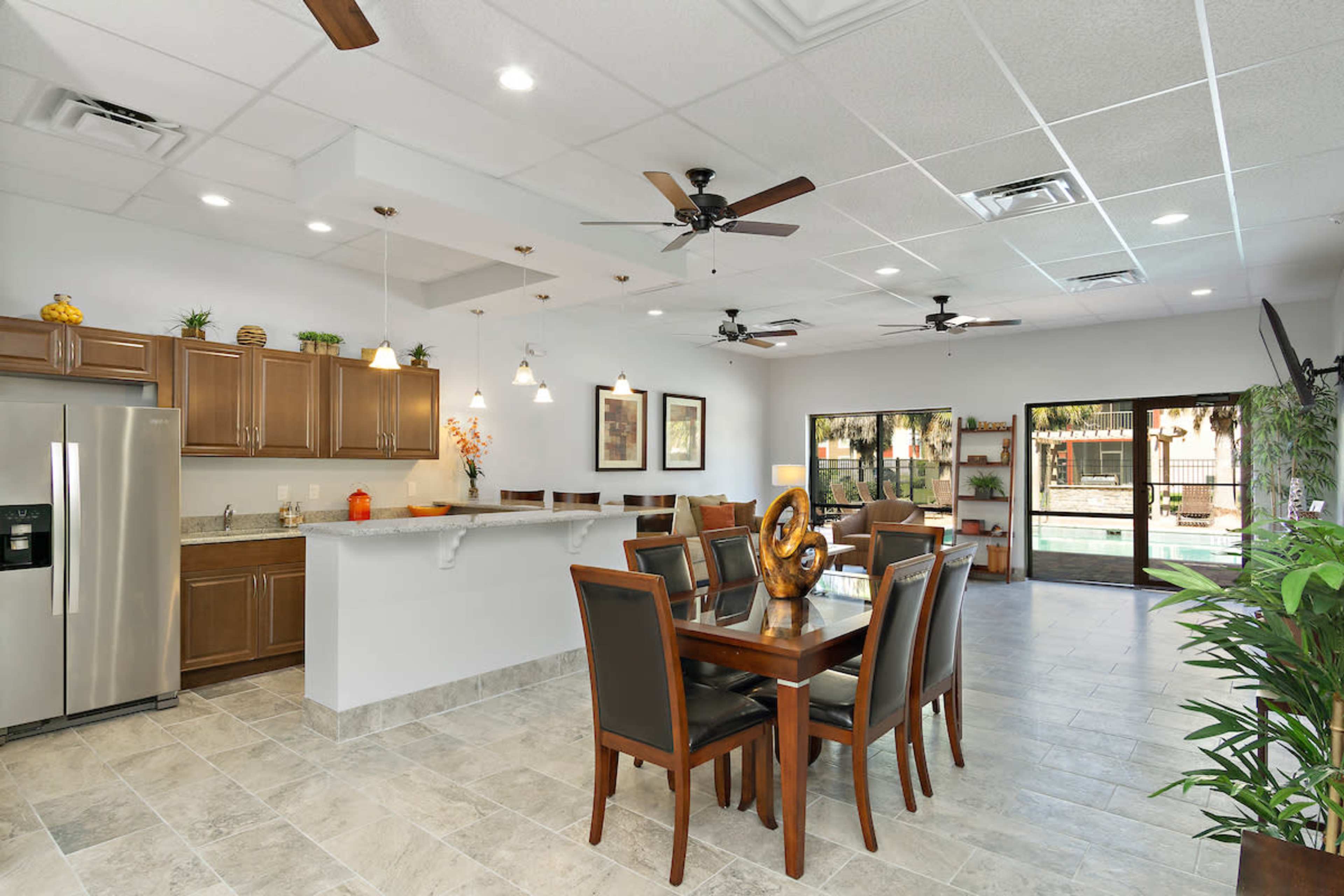 A spacious dining area features a large wooden table with chairs, a kitchen with dark cabinetry, and large windows that overlook a pool area.