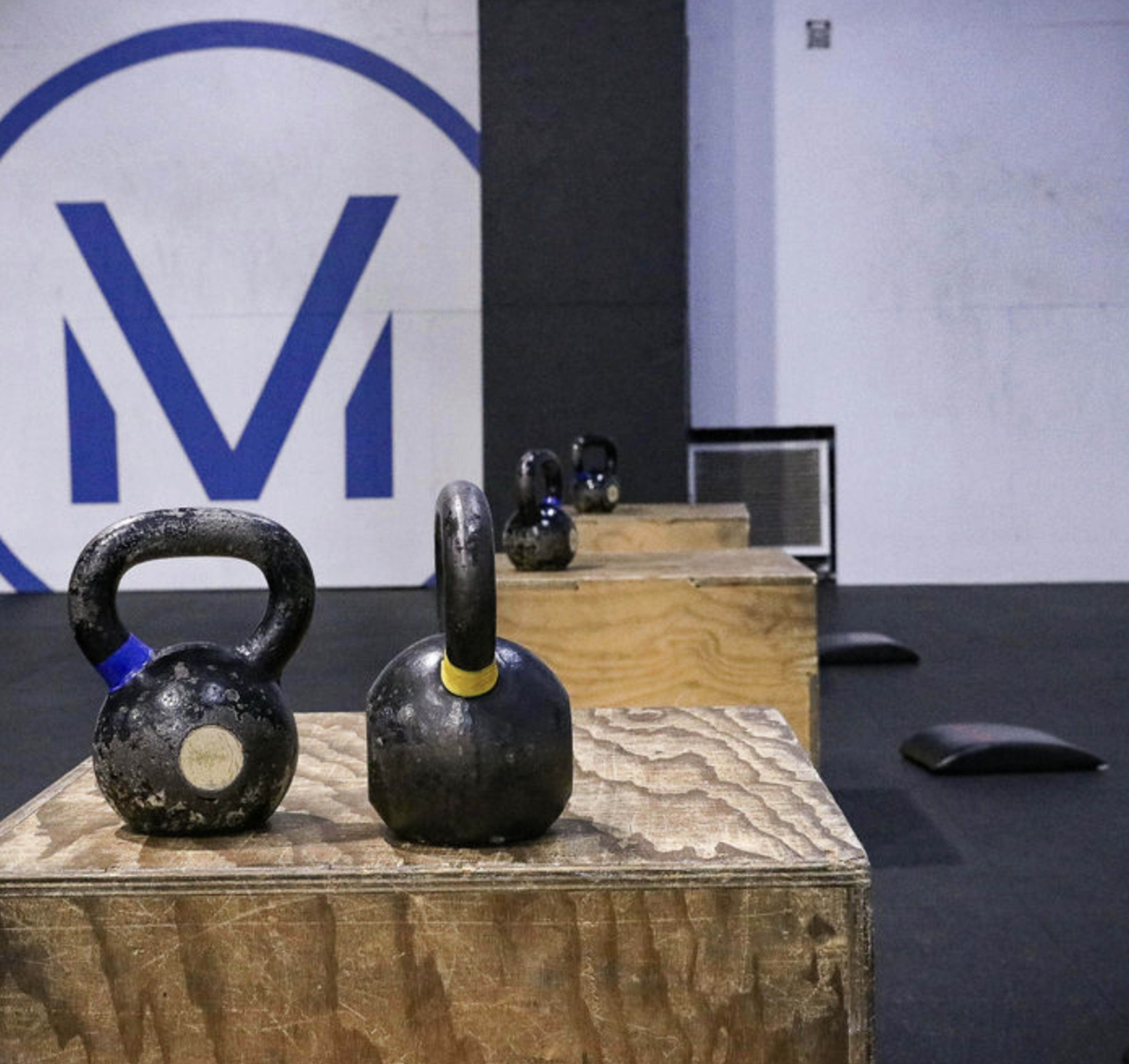 The image features a gym setting with several kettlebells placed on wooden boxes, arranged in a row against a wall with a large “M” logo.