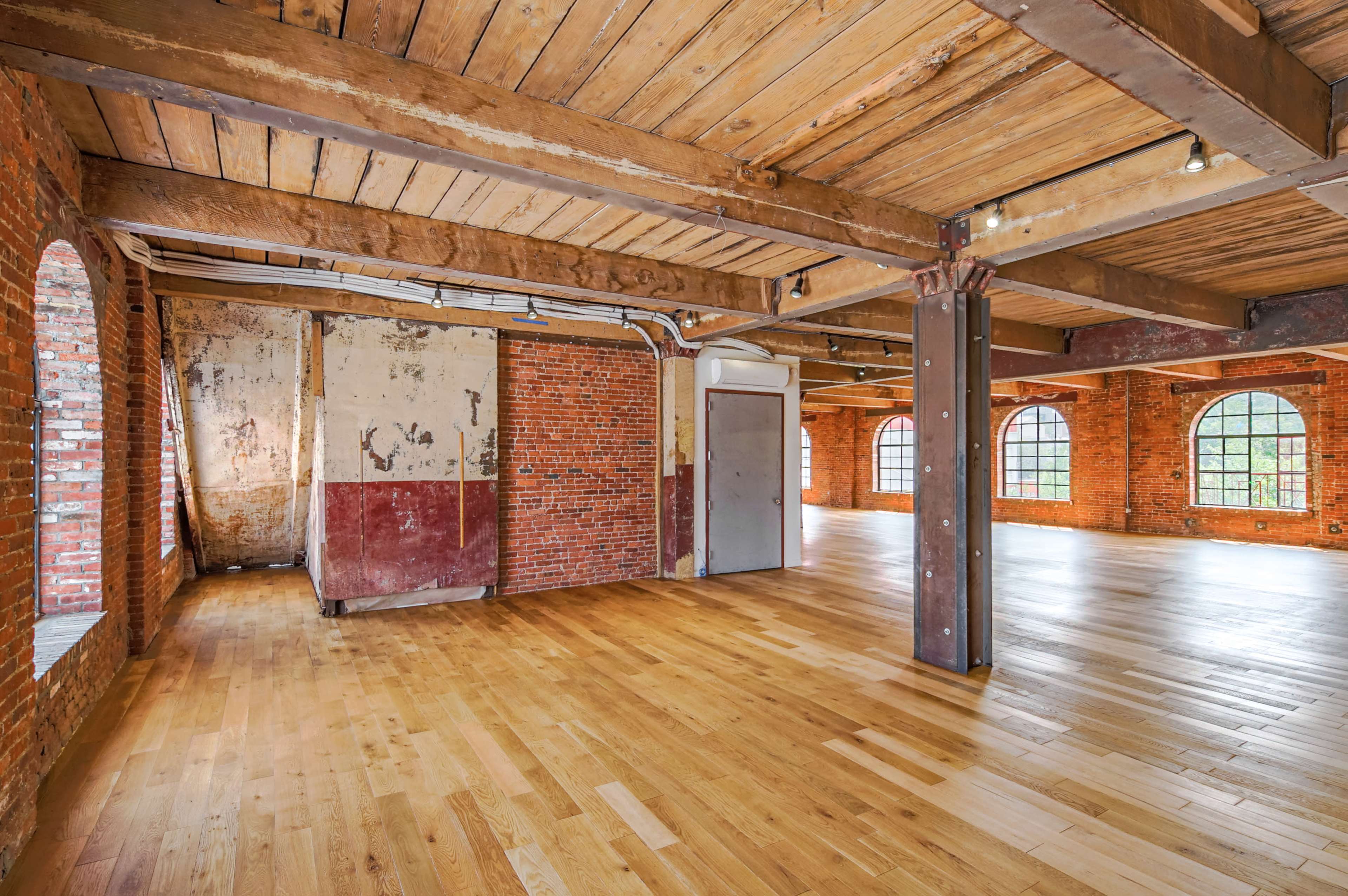 An open interior space features exposed brick walls, wooden beams, and a hardwood floor, with large arched windows allowing natural light.