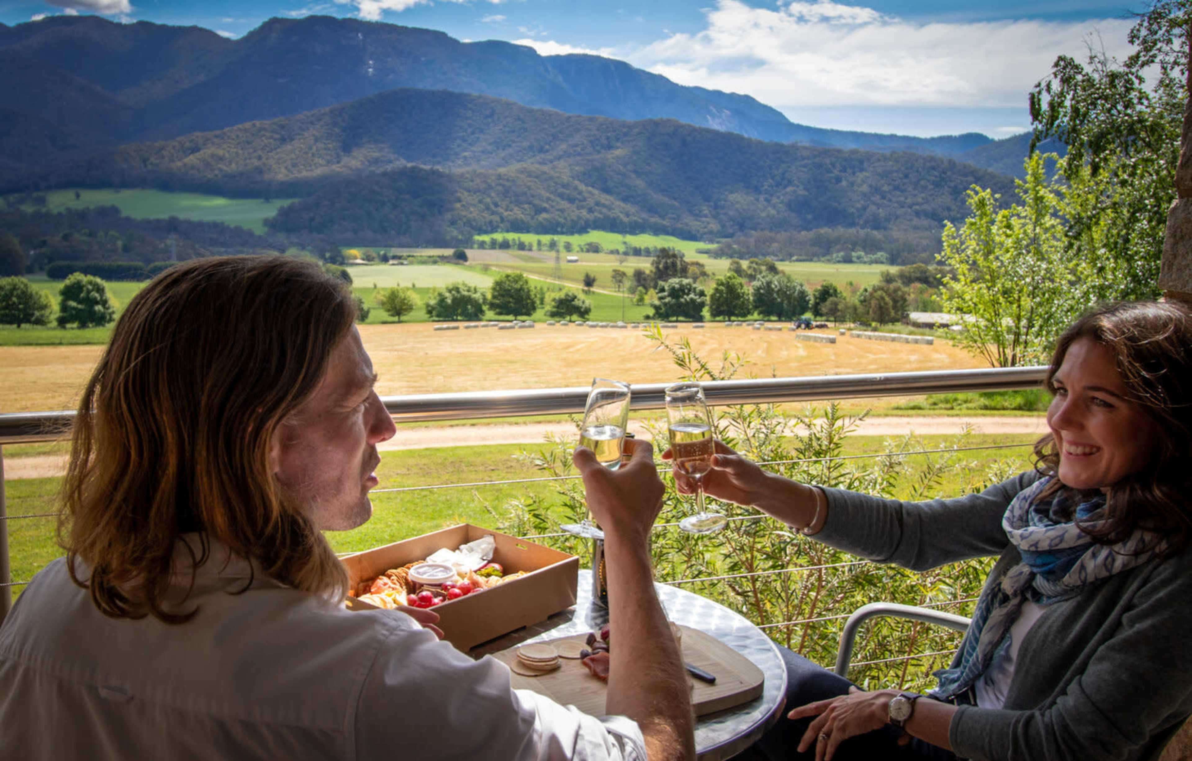 Two people toast with glasses of champagne while sitting at a table with a view of a mountain landscape.