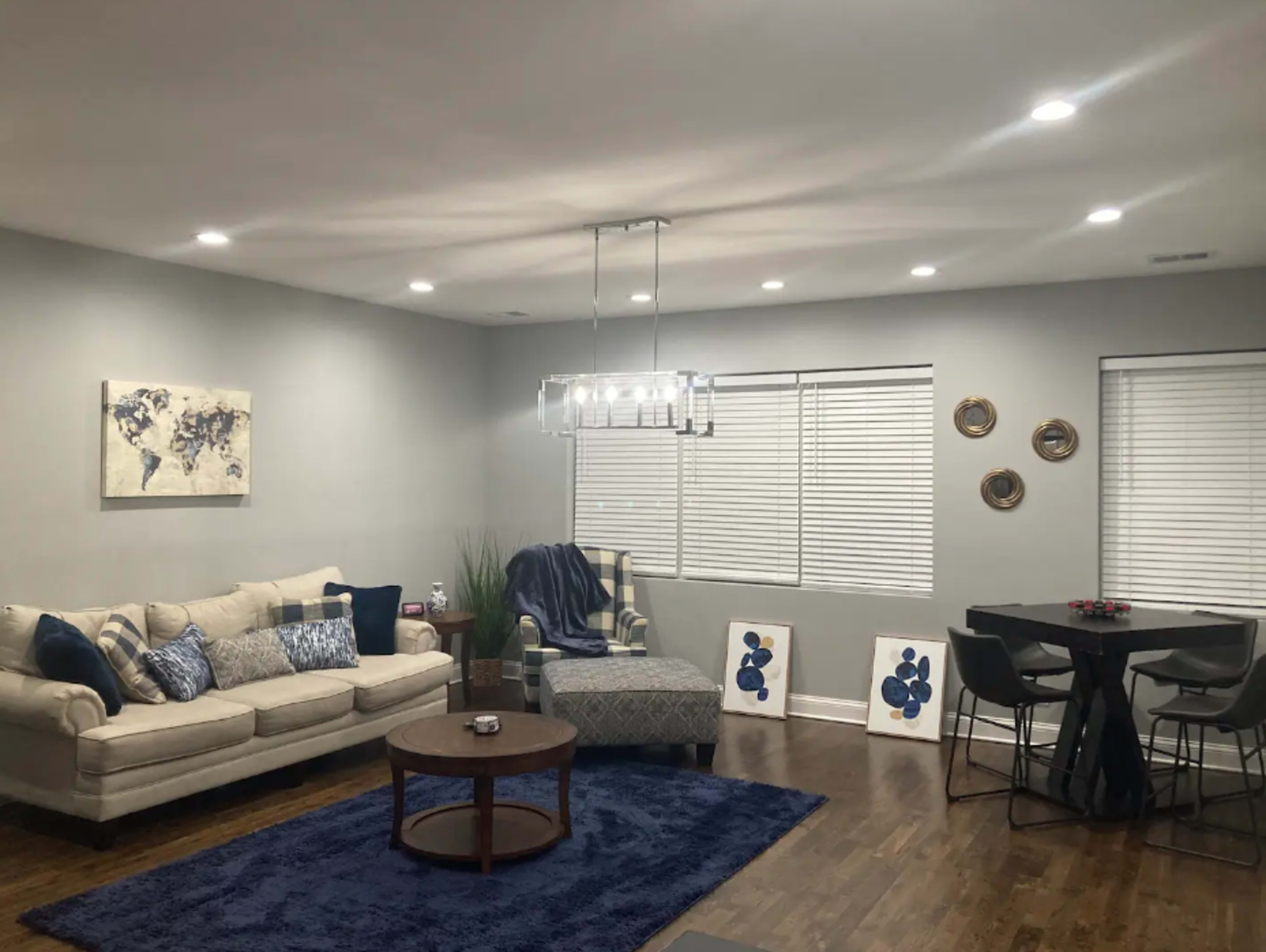 A living room features a beige sofa, a round coffee table, a dining area with black chairs, and wall decorations including framed art, all under bright ceiling lights.