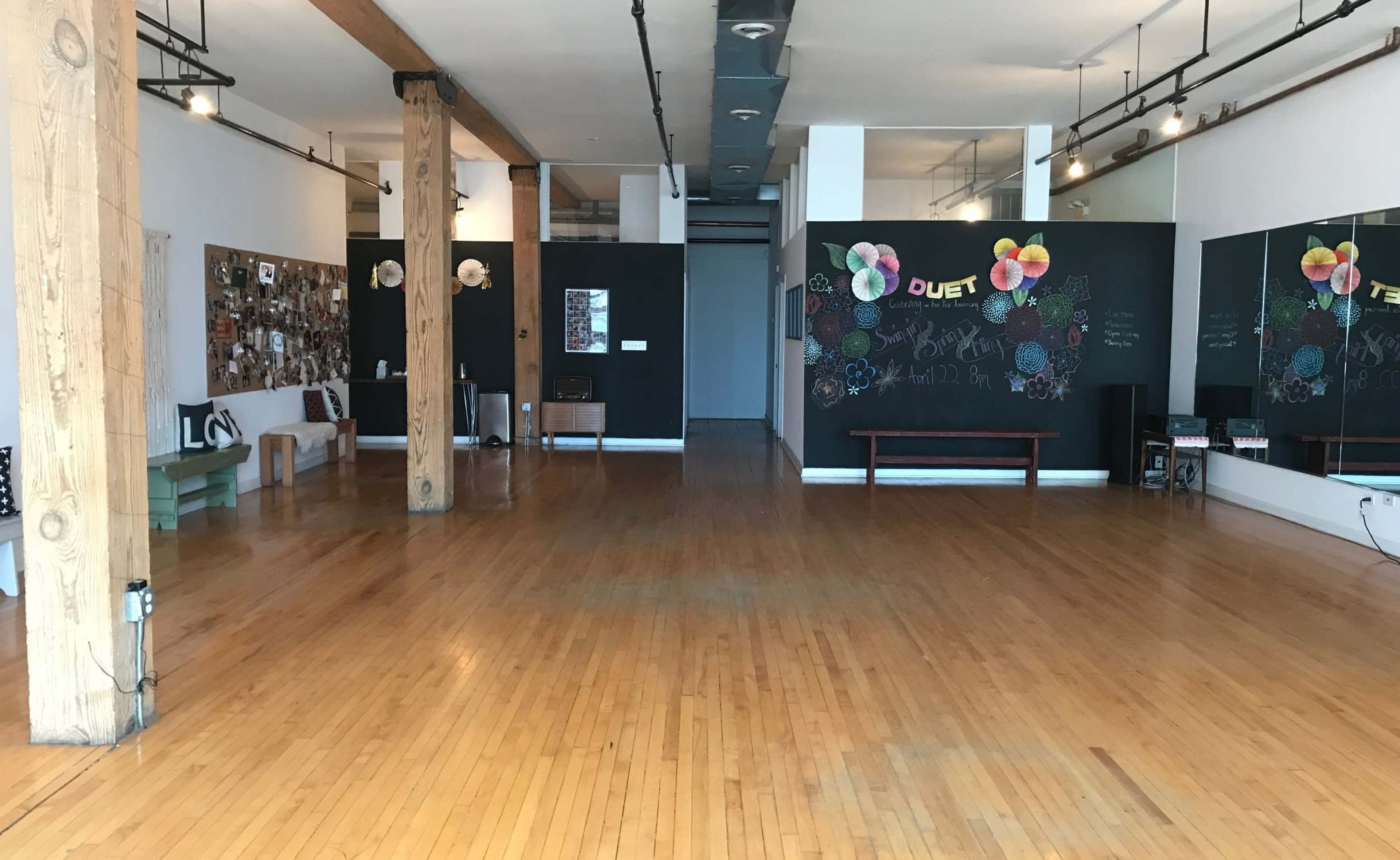 The image shows a spacious, empty studio with wooden floors, large mirrors on one wall, and black-painted walls decorated with colorful chalk art and photographs.
