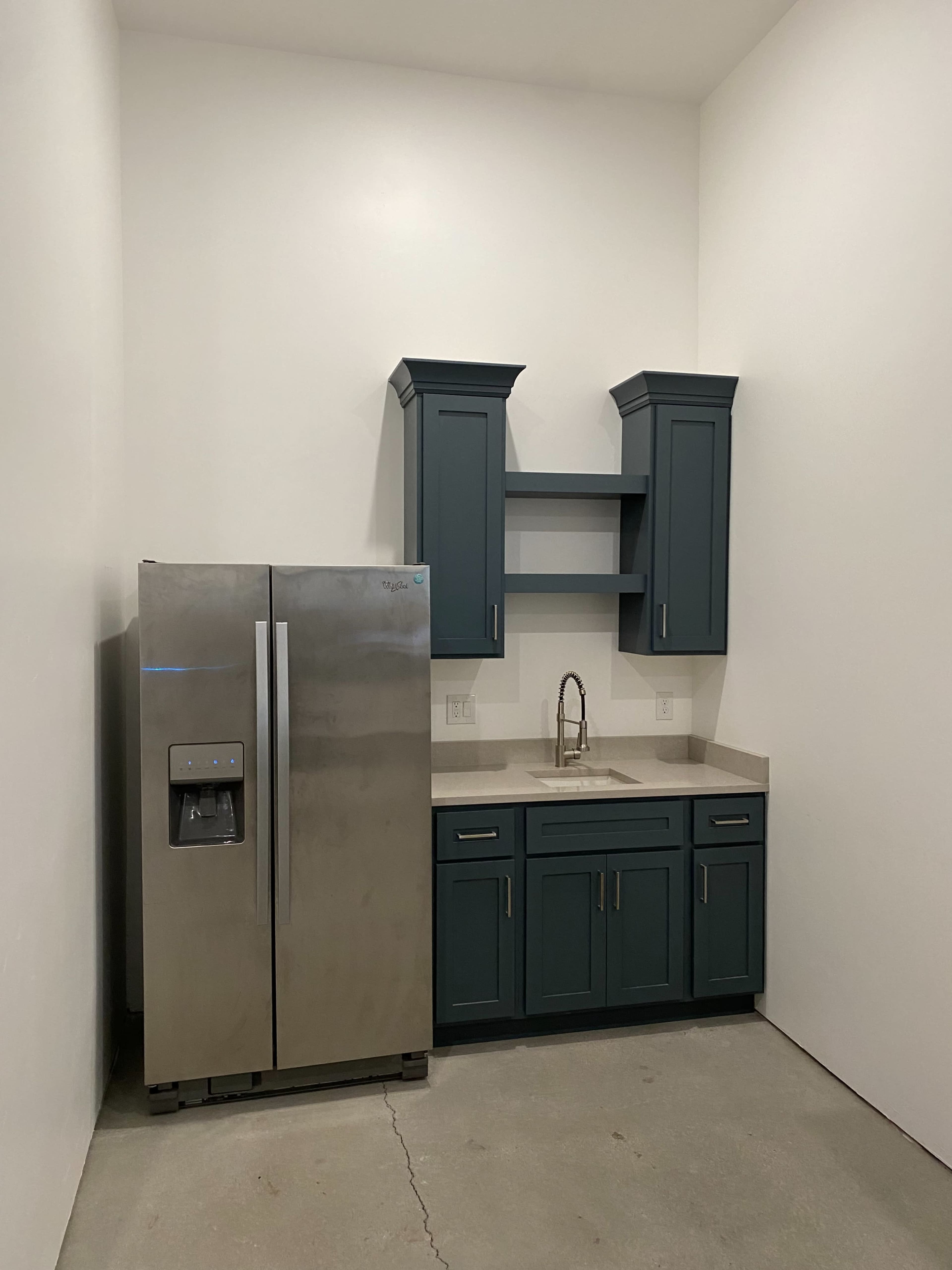 A kitchen corner features a stainless steel refrigerator beside dark blue cabinets and a sink with a countertop.
