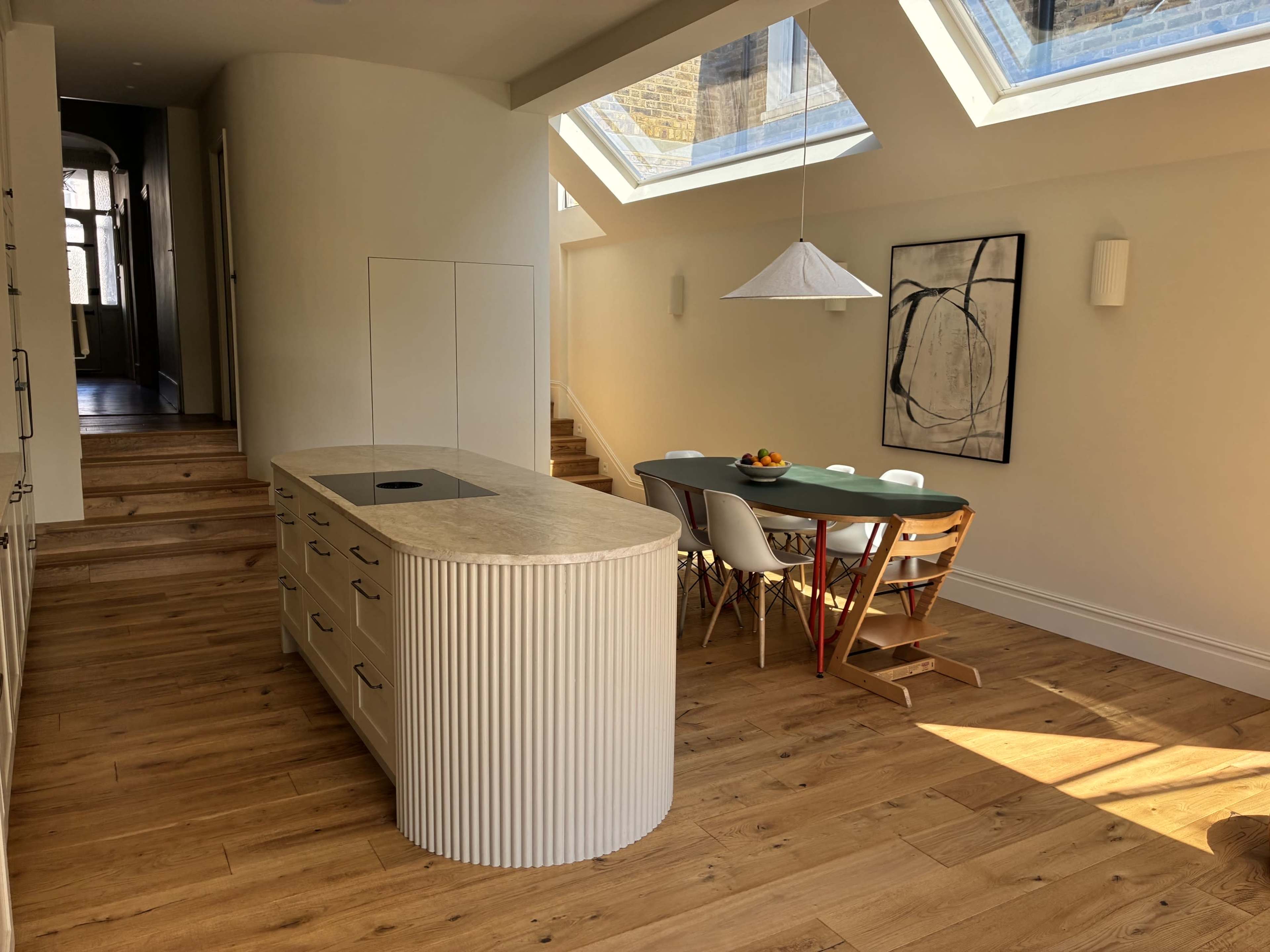 The image shows a modern kitchen with a light-colored island and dining area featuring a green table and various chairs, illuminated by skylights.