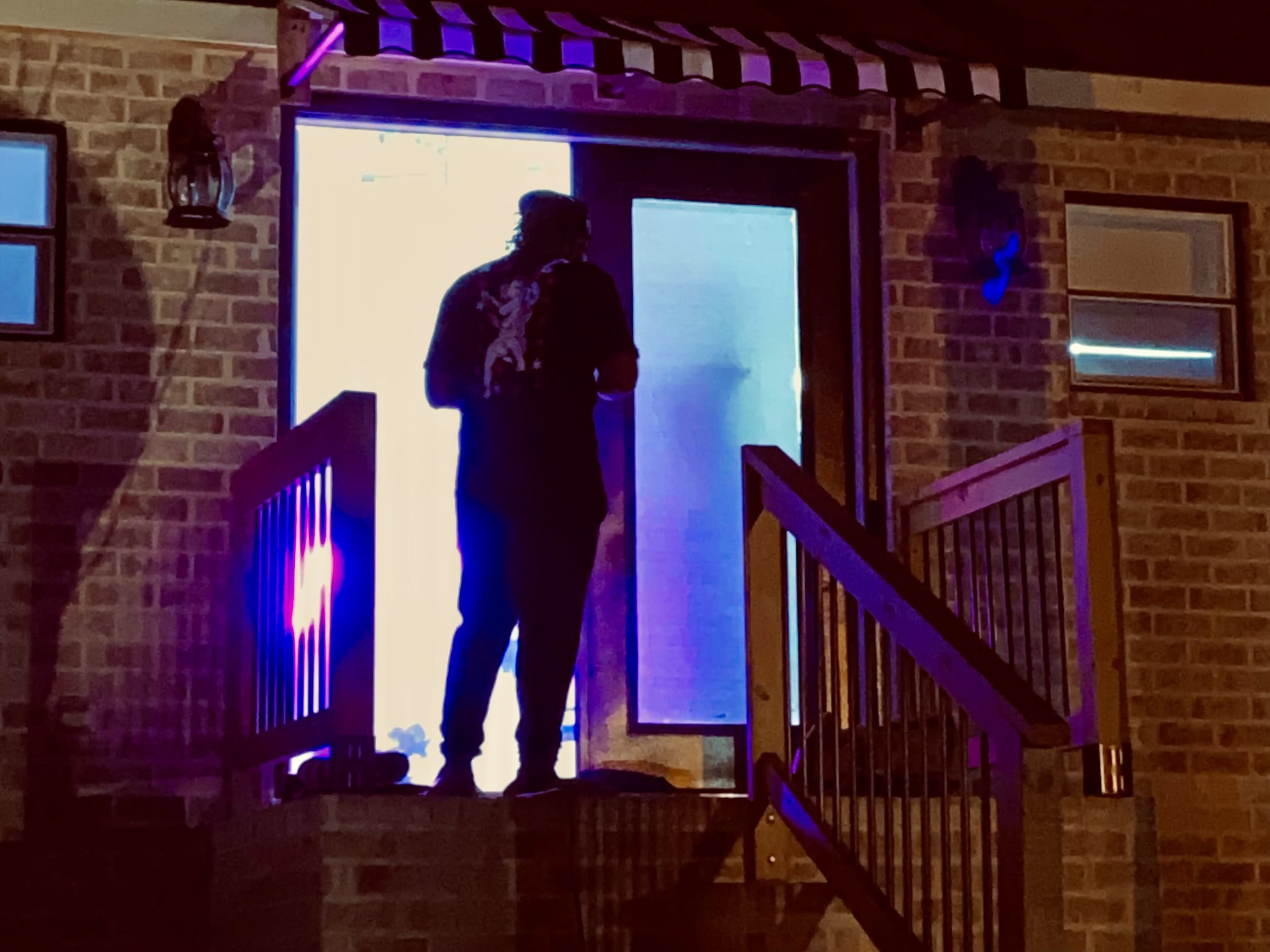 A person stands on the steps of a house, silhouetted against a brightly lit doorway.
