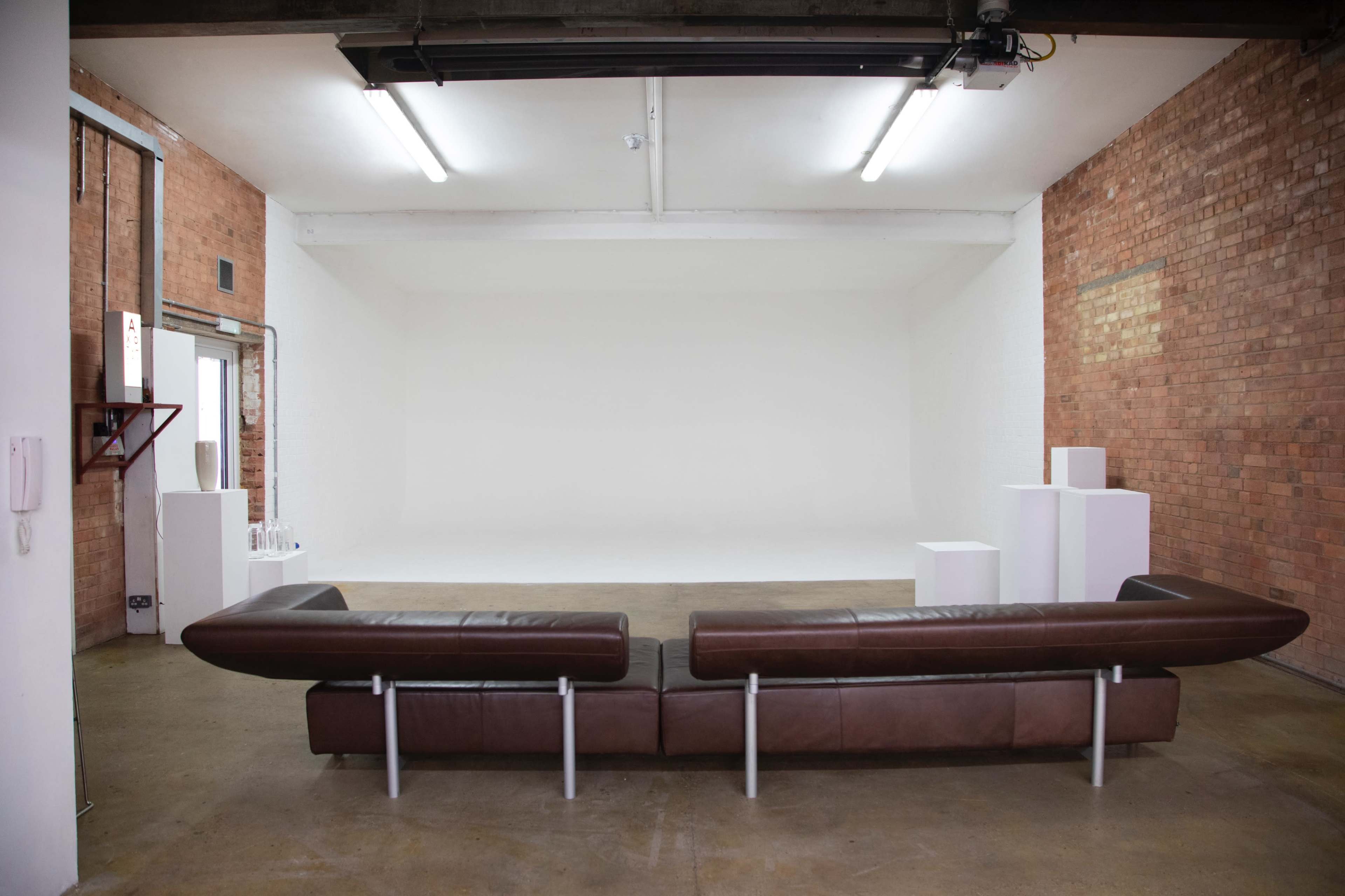 An empty studio space with a brown leather sofa facing a plain white wall, surrounded by brick walls and a polished concrete floor.