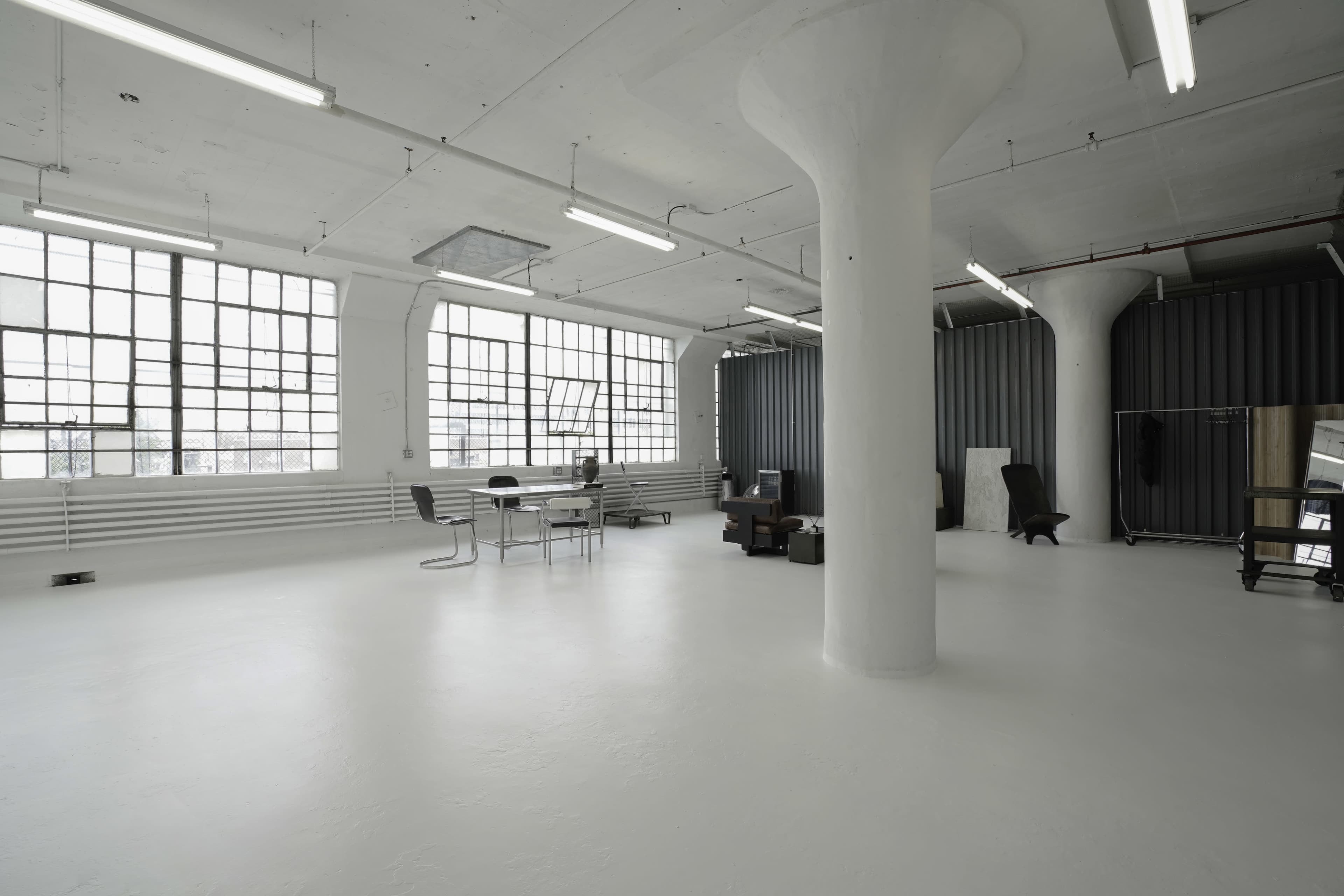 A spacious, minimalist room with concrete pillars, large windows, and sparse furniture including a table, chairs, and a clothing rack.