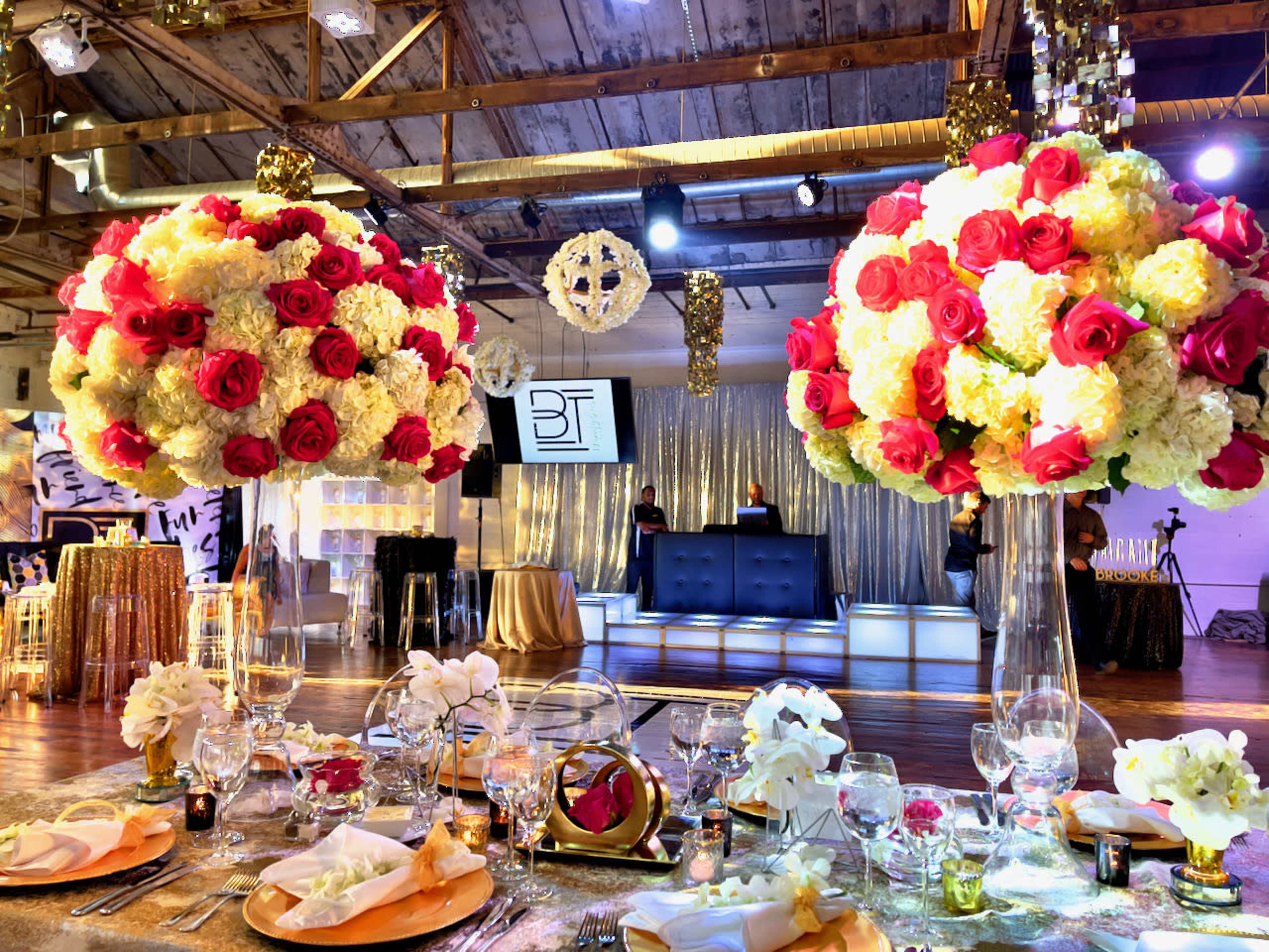 A wedding reception setup with large floral centerpieces of roses and hydrangeas, elegantly decorated tables, and a DJ booth in the background.