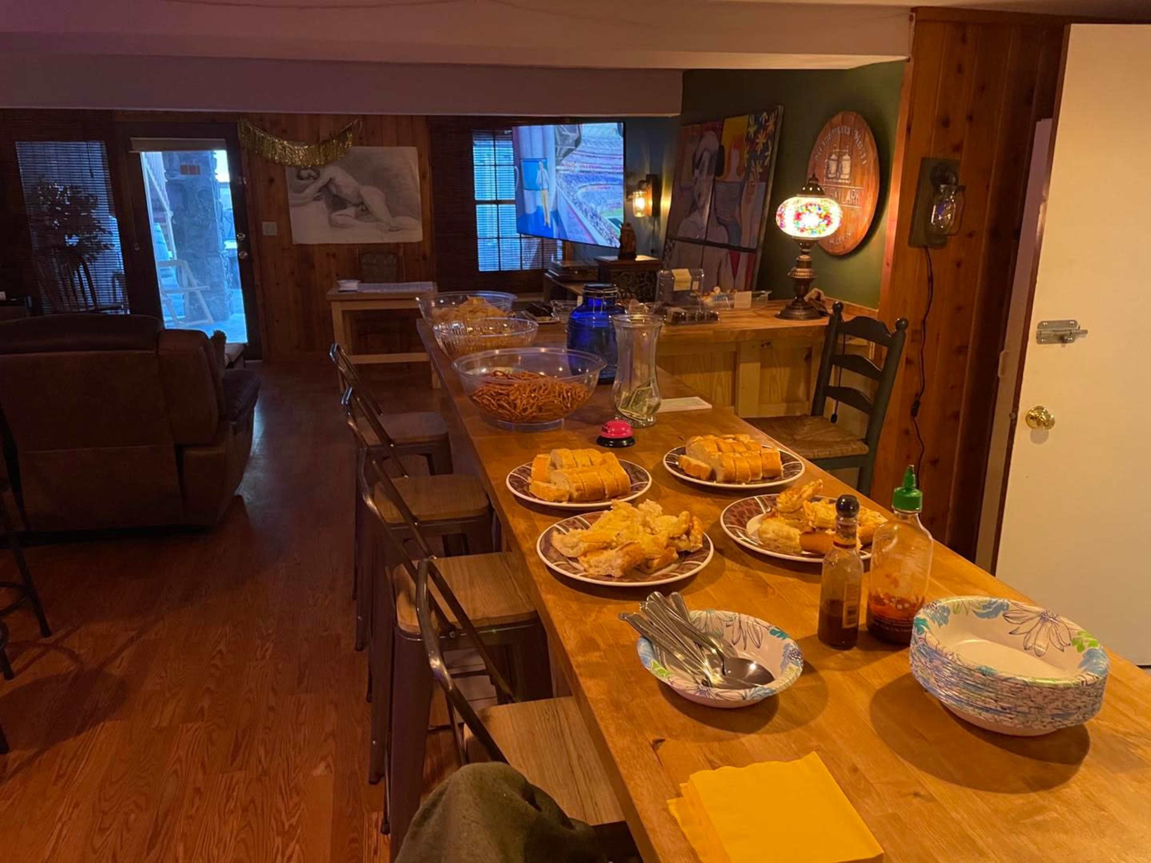 A dining table is set with various plates of food and condiments in a cozy wooden interior, with a view of a living area in the background.