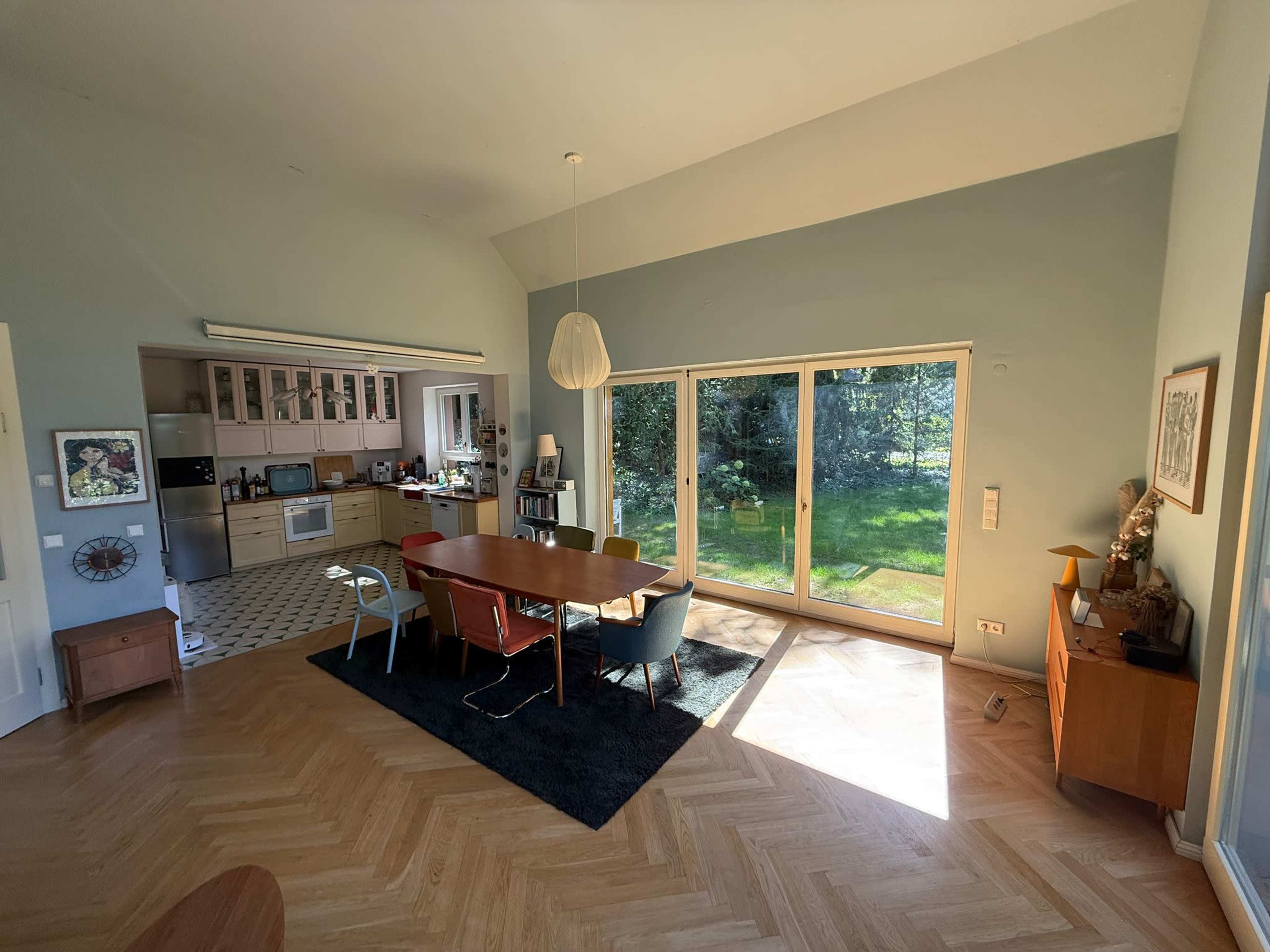 A spacious living area features a dining table surrounded by chairs, with large windows offering a view of a green outdoor space.