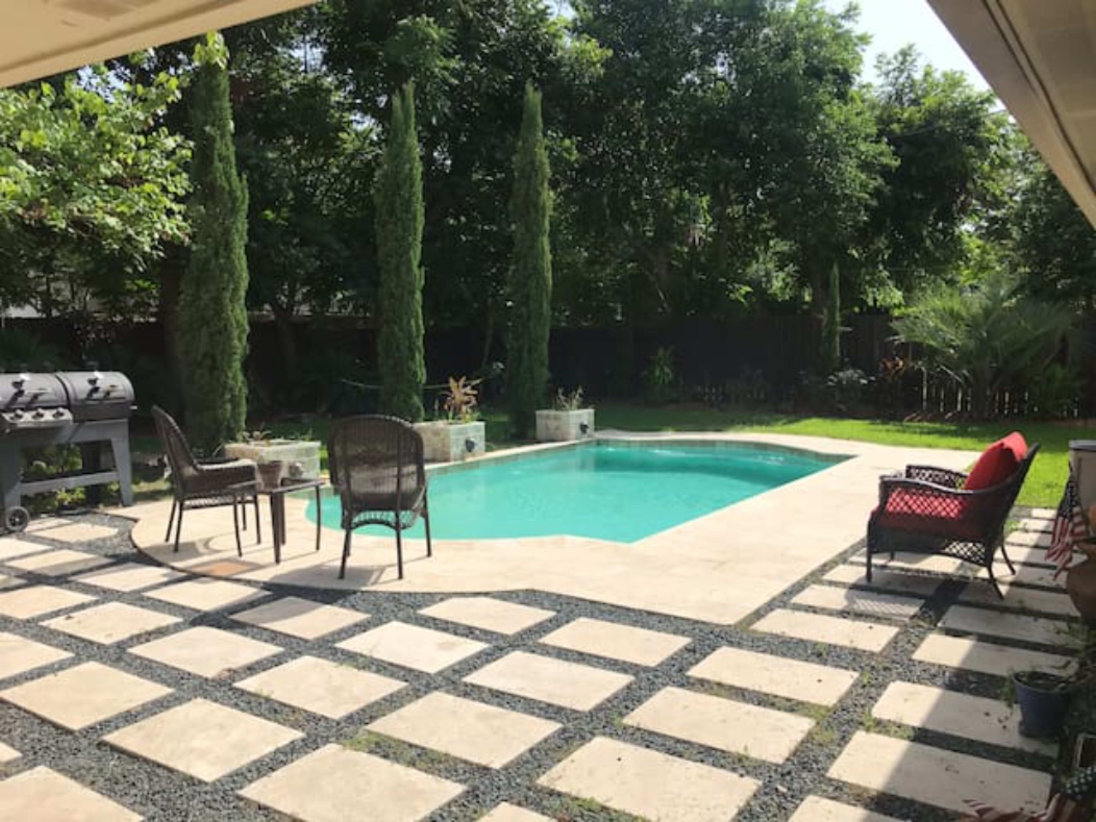 A backyard with a swimming pool surrounded by tall trees, stone pavers, and outdoor furniture.
