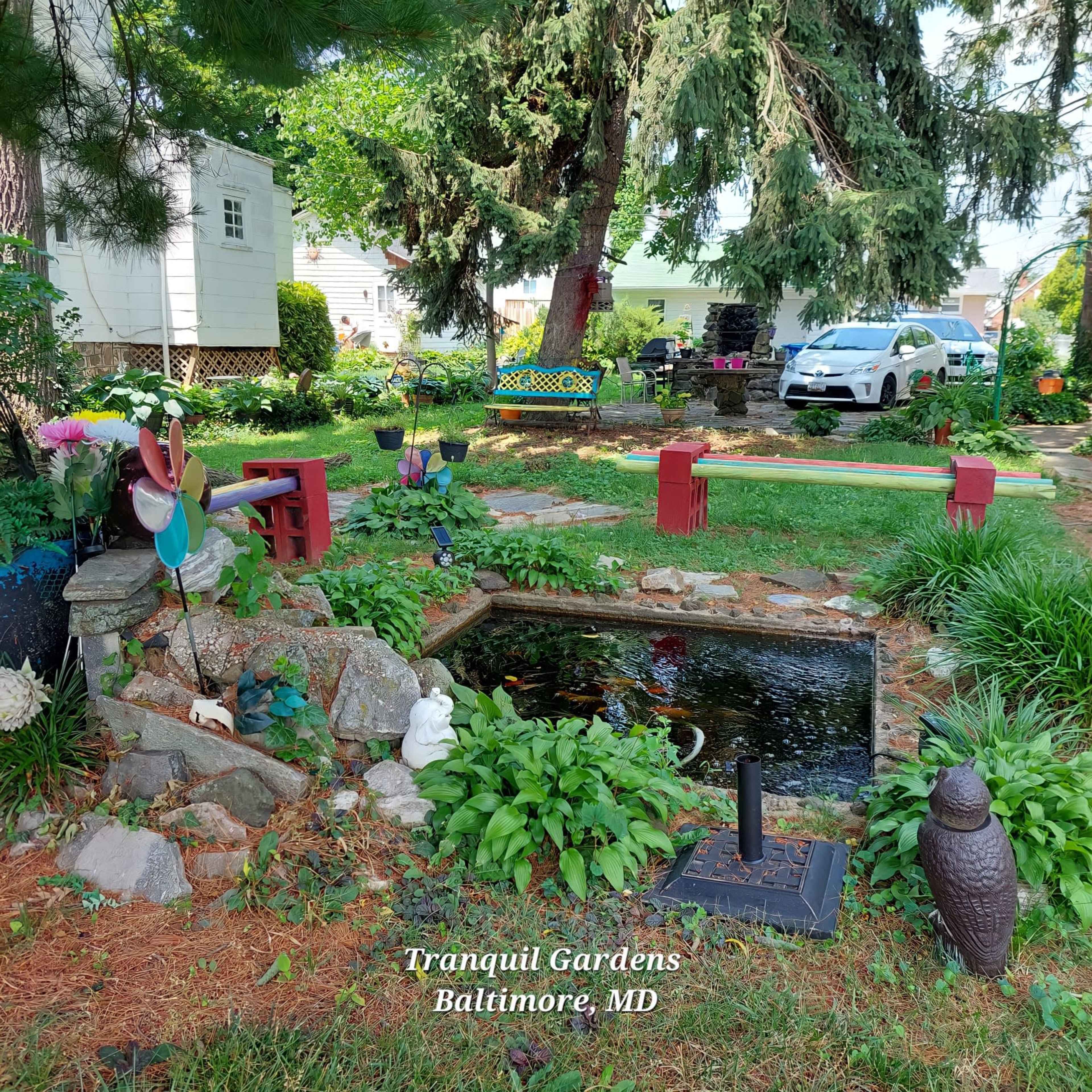 The image shows a landscaped garden in Baltimore, MD, featuring a pond surrounded by plants, decorative stones, and various seating areas.