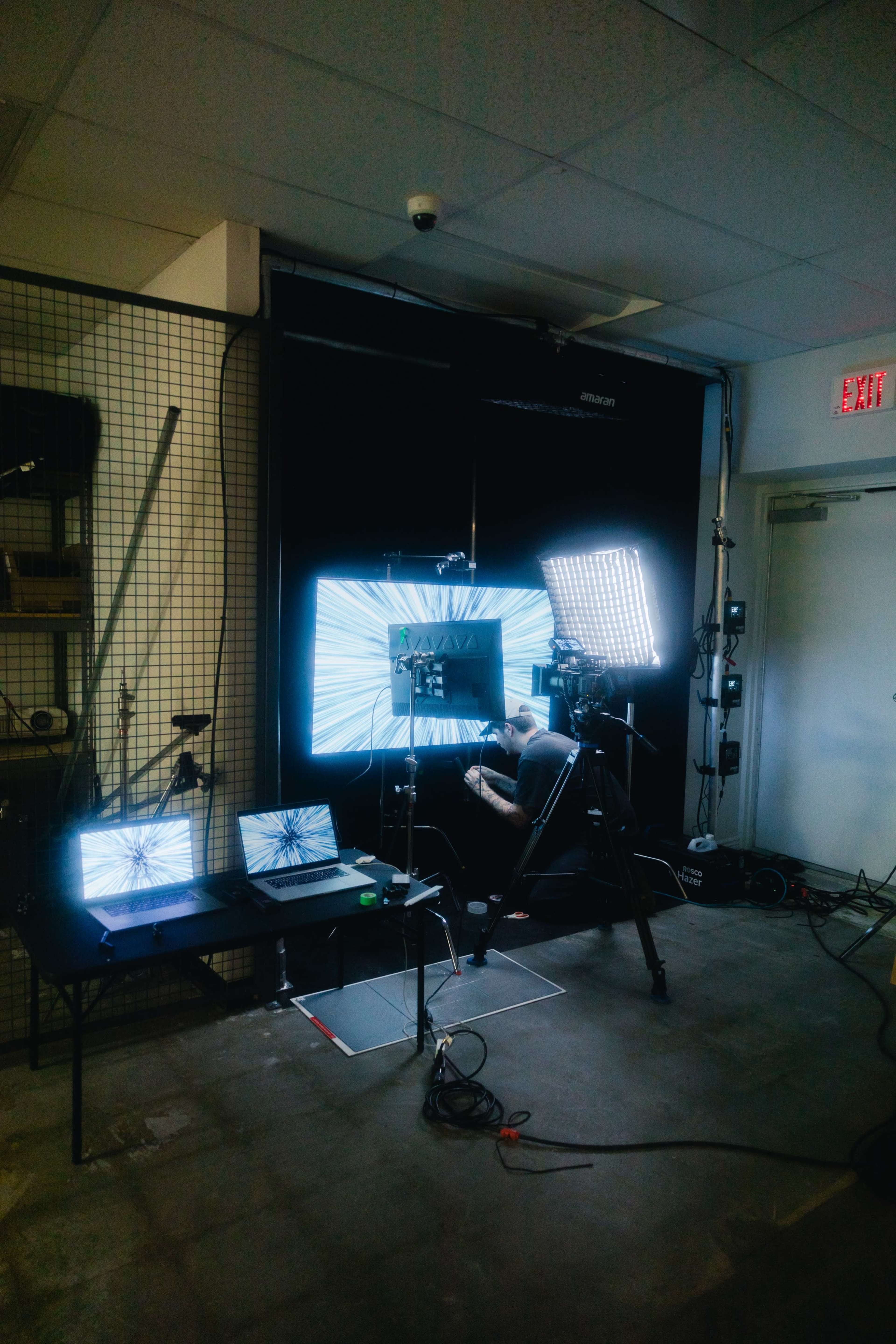 A person operates a camera and monitors in a studio illuminated by bright lights and a blackout backdrop.