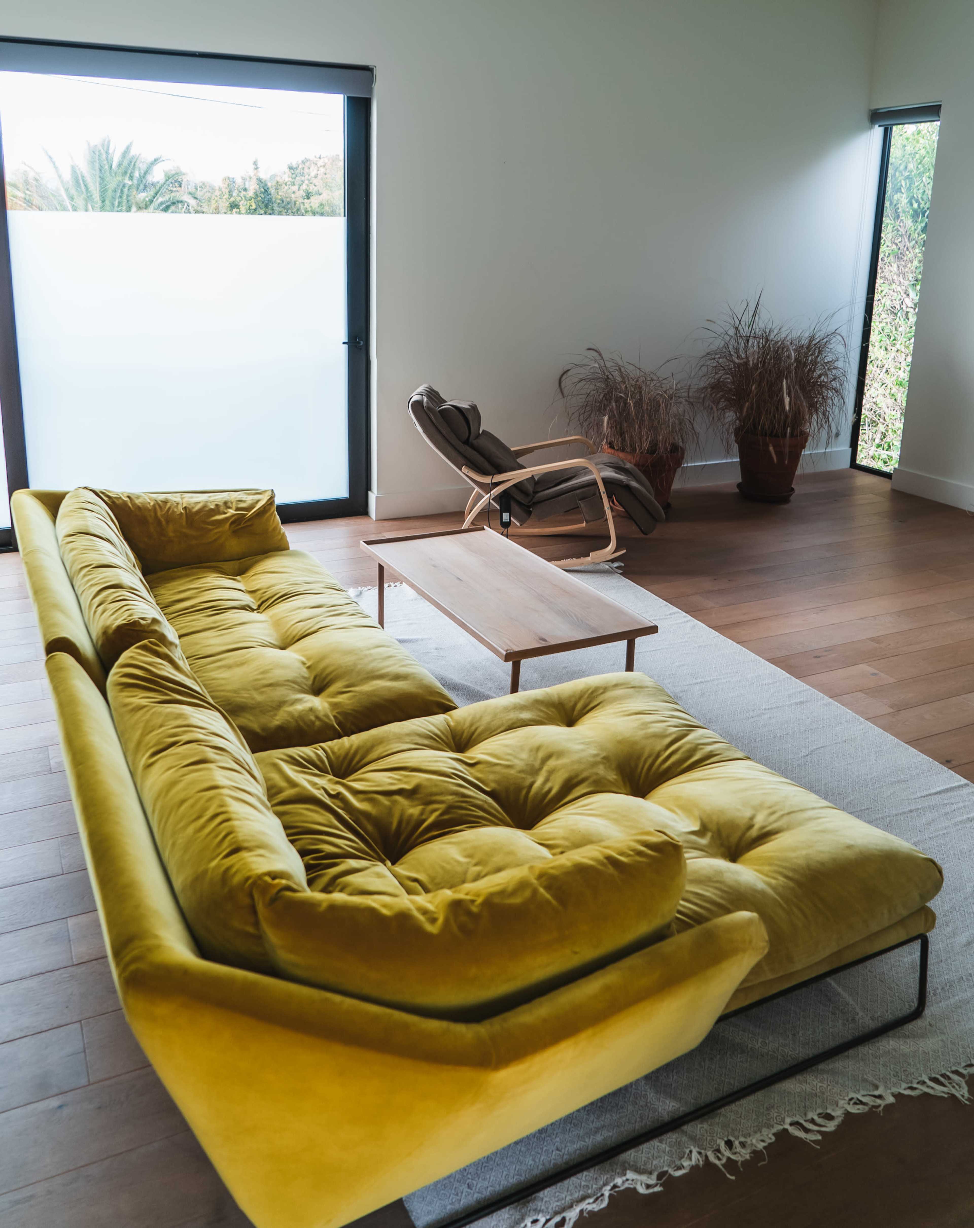 A yellow sectional sofa faces a wooden coffee table in a modern living room with large windows overlooking greenery.