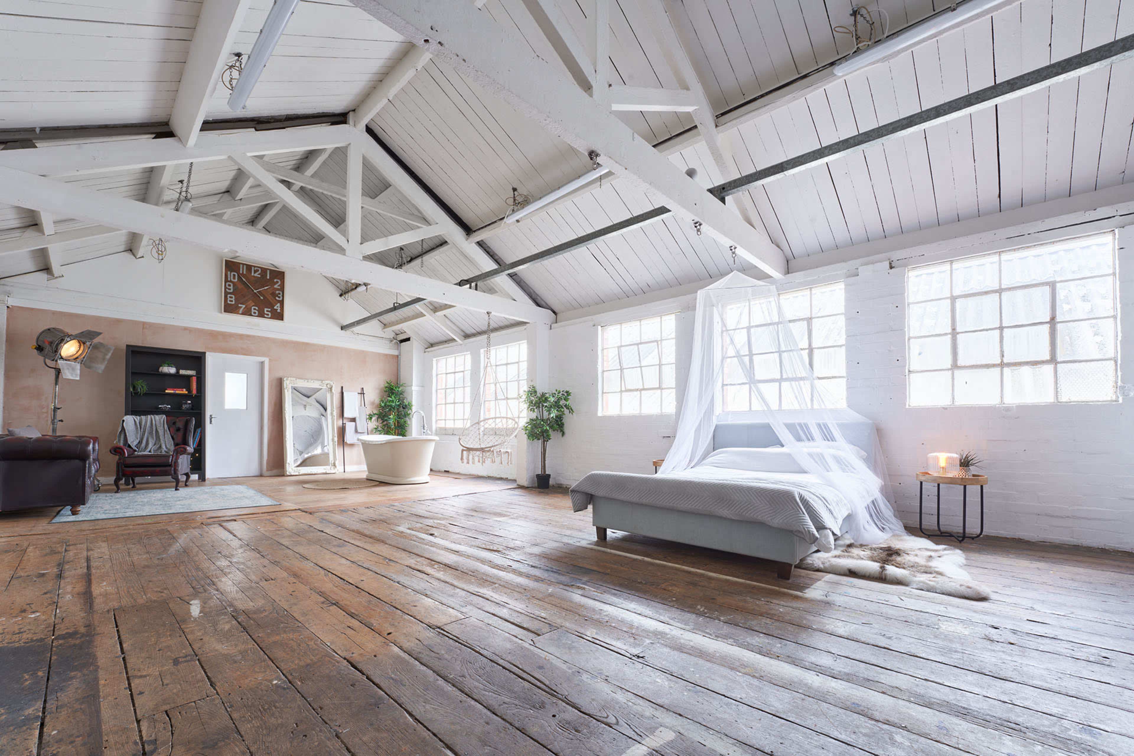 A spacious loft-style bedroom features a bed with a sheer canopy, large windows, and exposed wooden beams, with a freestanding bathtub and a cozy sitting area in the background.