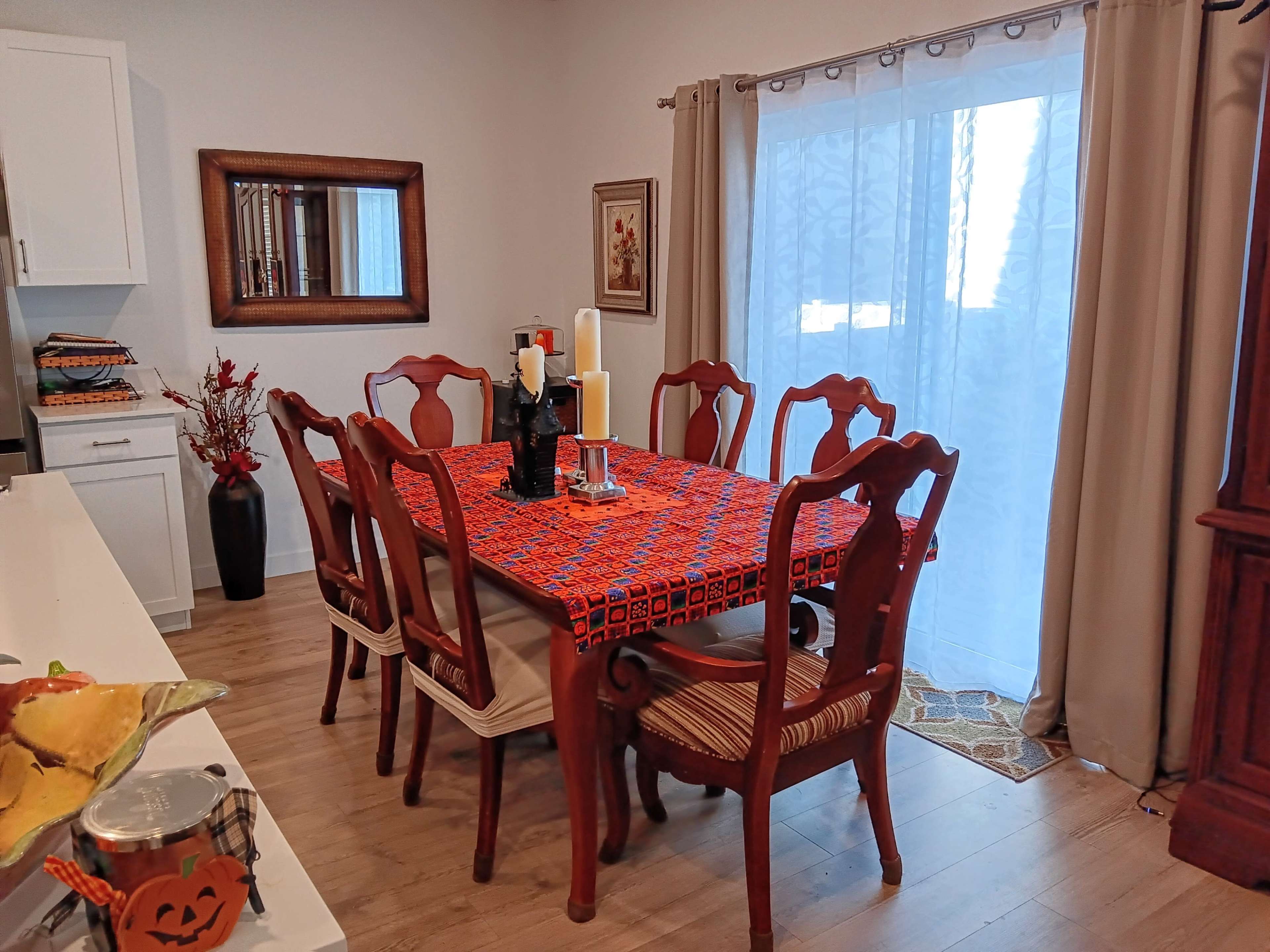 A dining room features a wooden table covered with an orange tablecloth and surrounded by six chairs, with a window draped in sheer curtains.