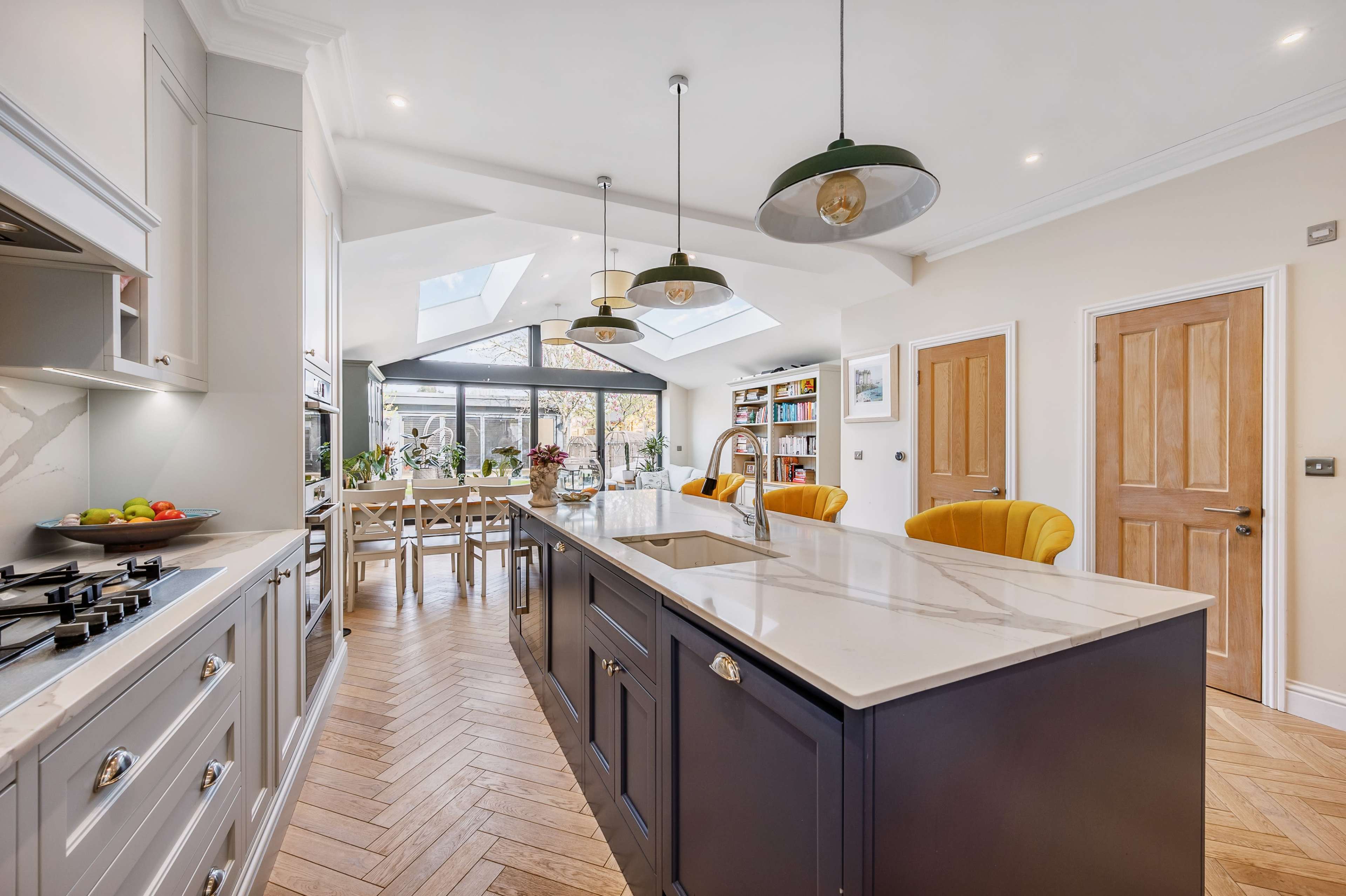 A modern kitchen features a central island with a marble countertop, surrounded by yellow chairs and connected to a bright dining area with large windows and shelves.