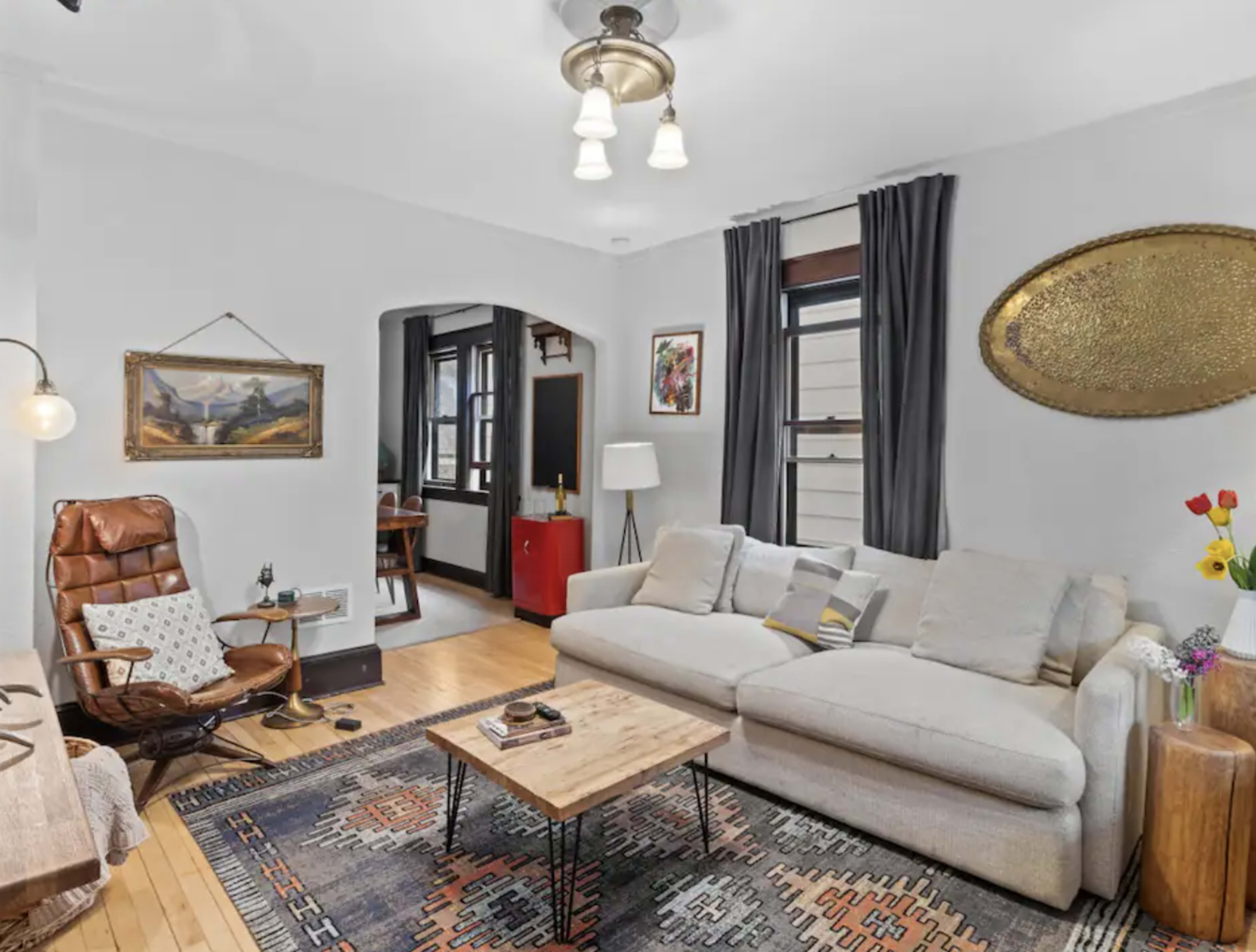 The image shows a cozy living room with a light-colored sofa, a wooden coffee table, an armchair, and artwork on the walls.