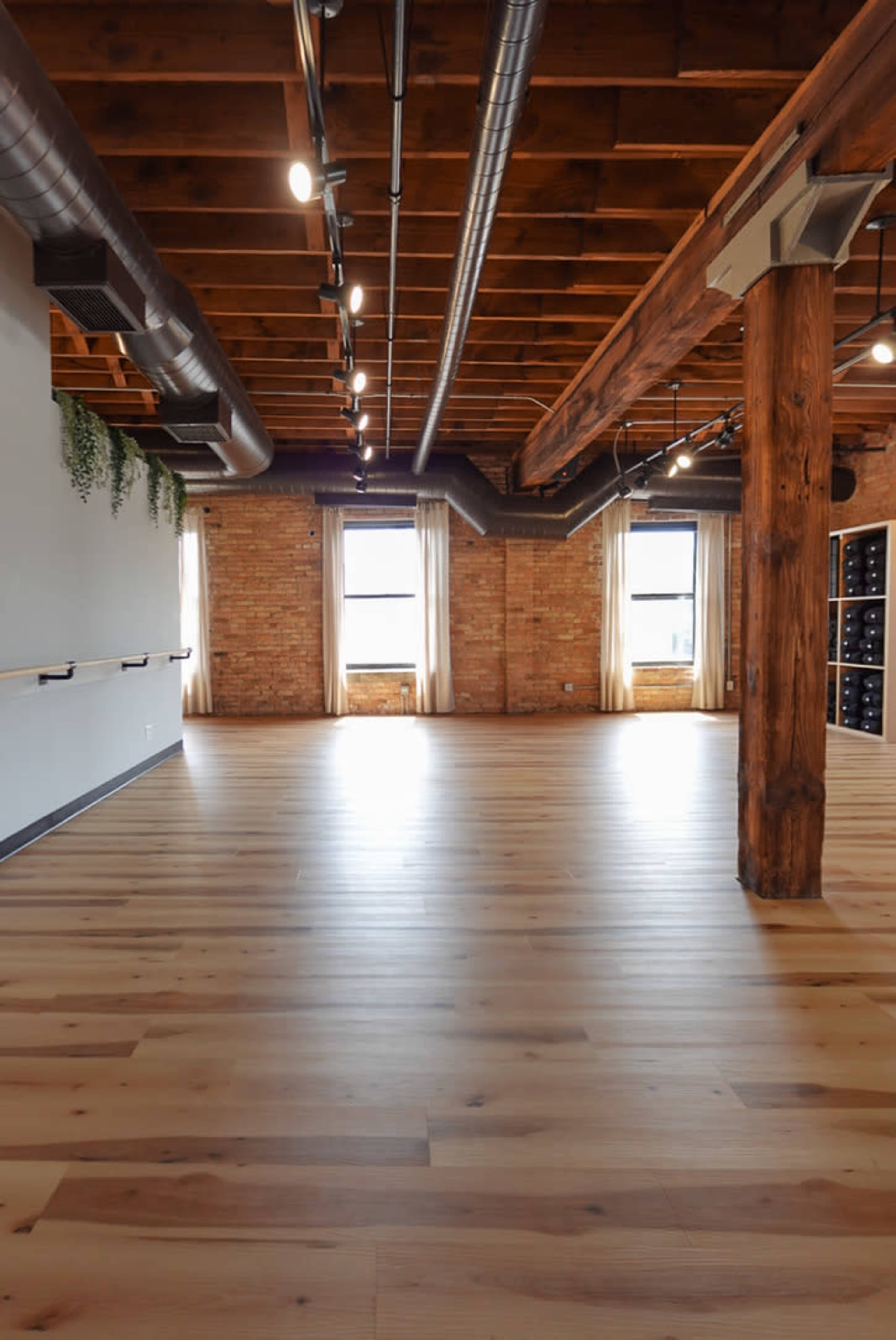 The image shows a spacious interior with hardwood floors, exposed wooden beams, and large windows, creating an open and airy atmosphere.