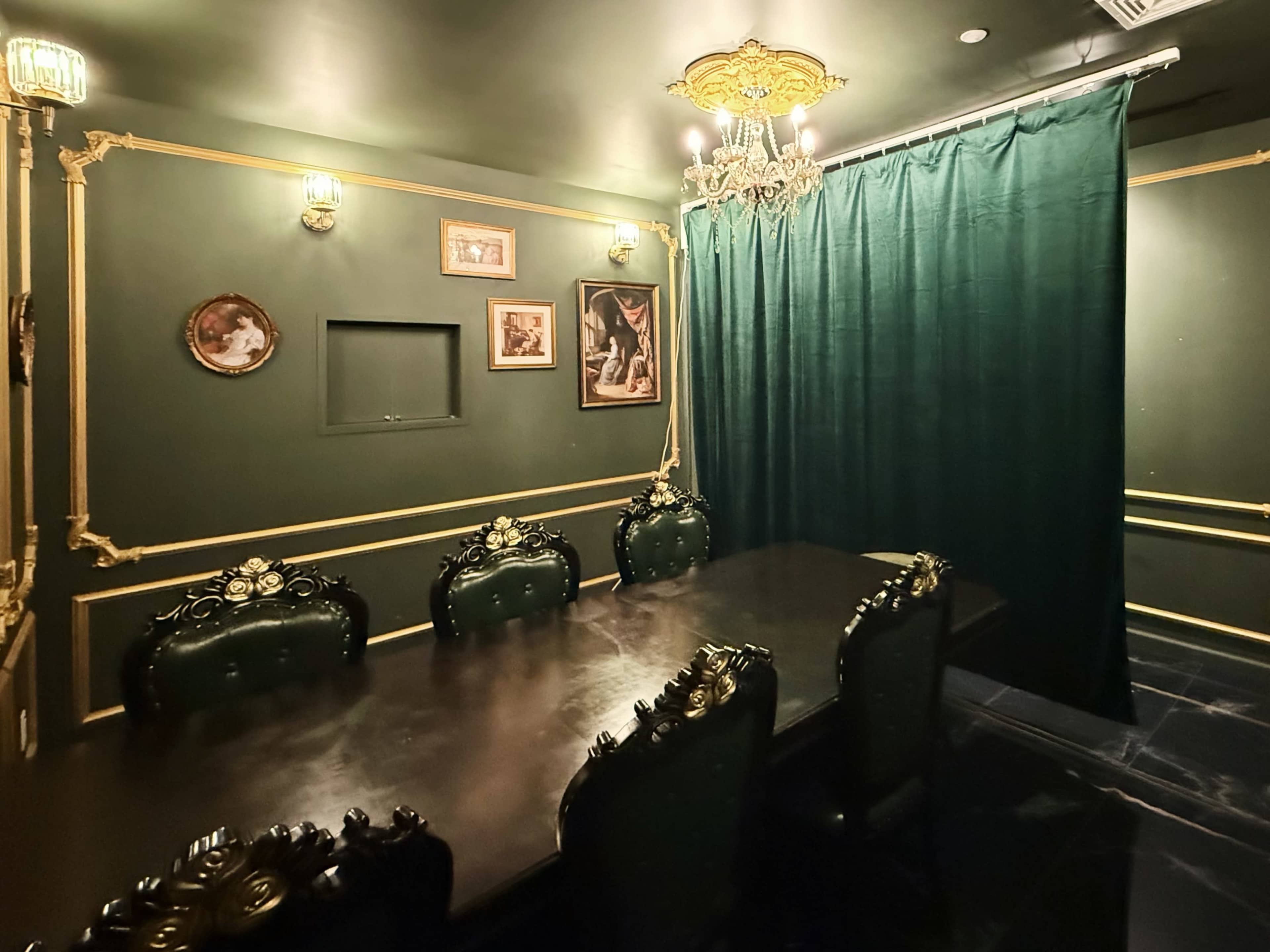 The room features a dark green decor with a large table surrounded by ornate chairs, framed artwork on the walls, and a curtain separating a section of the space.
