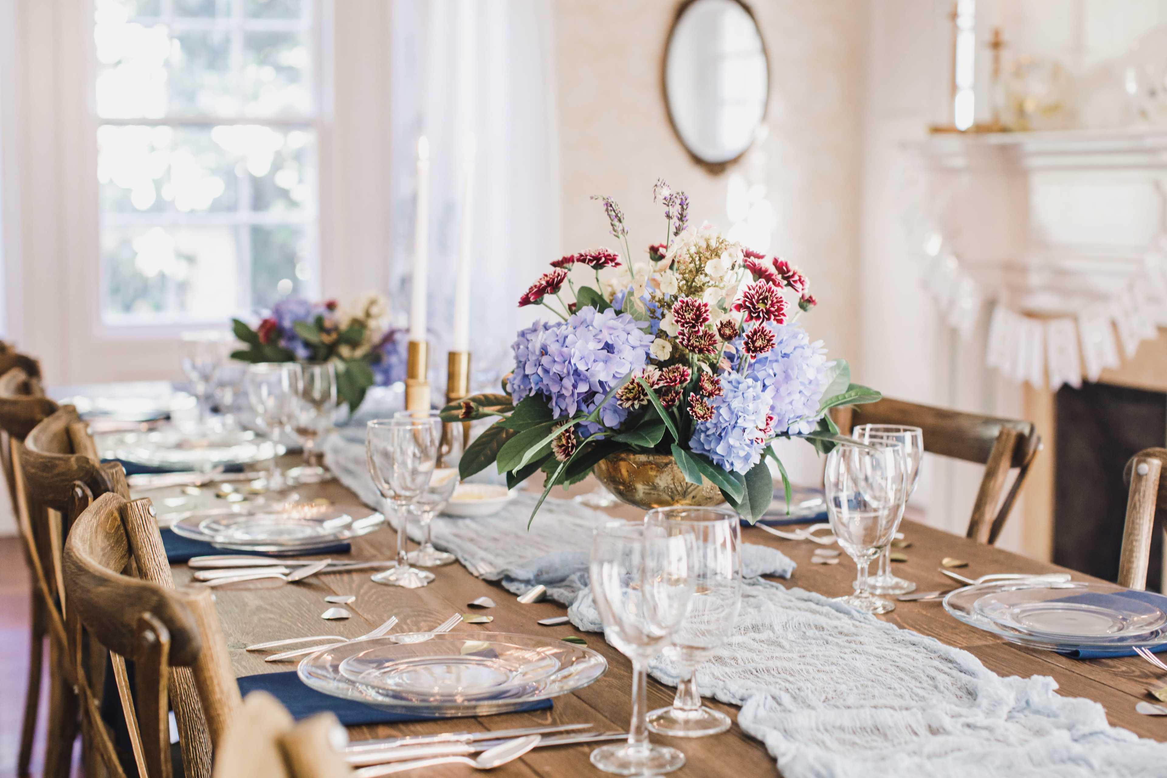 A wooden dining table is set with glasses, plates, and a floral centerpiece, featuring blue hydrangeas and assorted flowers.