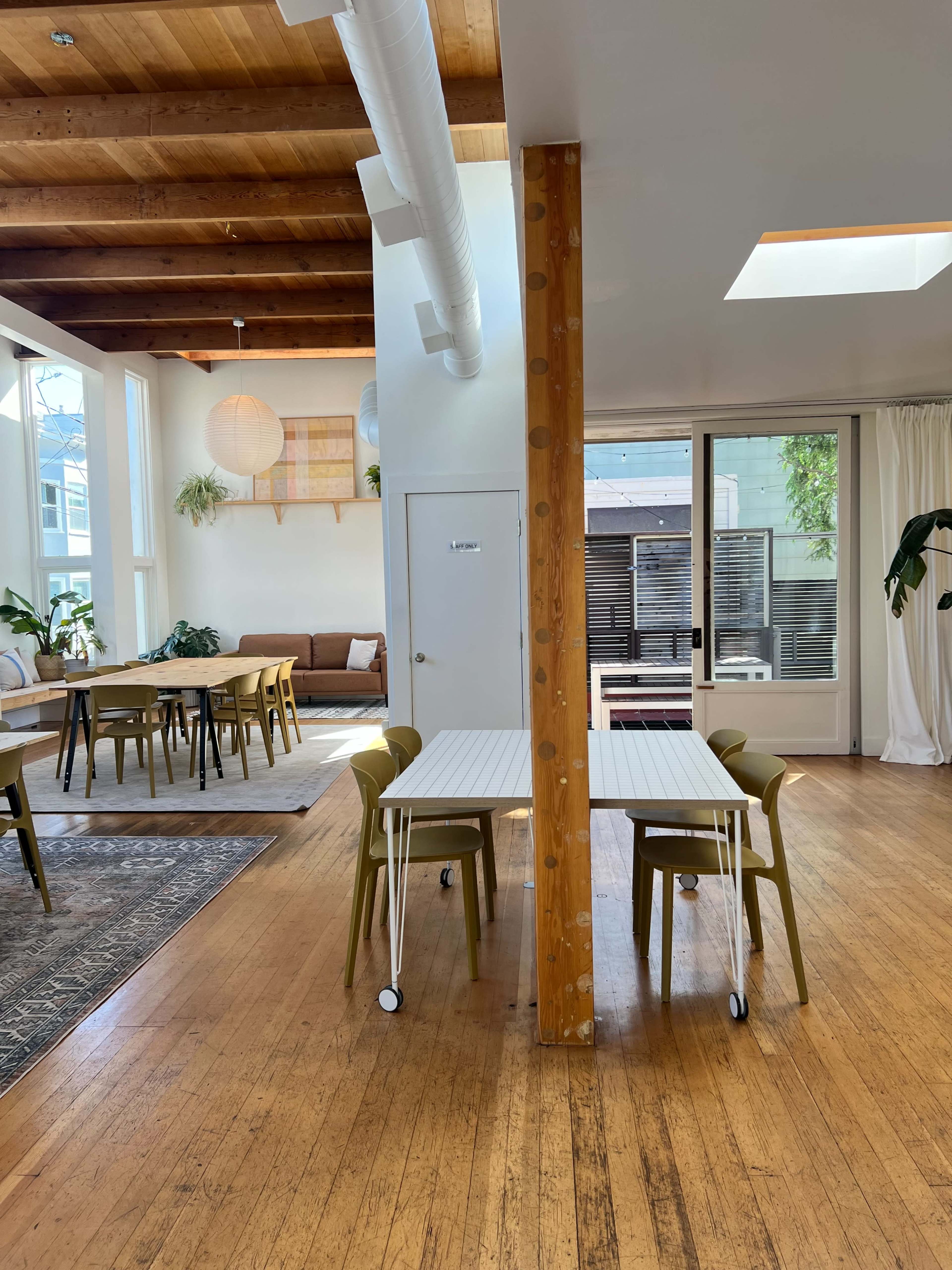 A spacious, well-lit room with wooden floors, featuring a divider post between two sets of tables and chairs, along with large windows and plants.