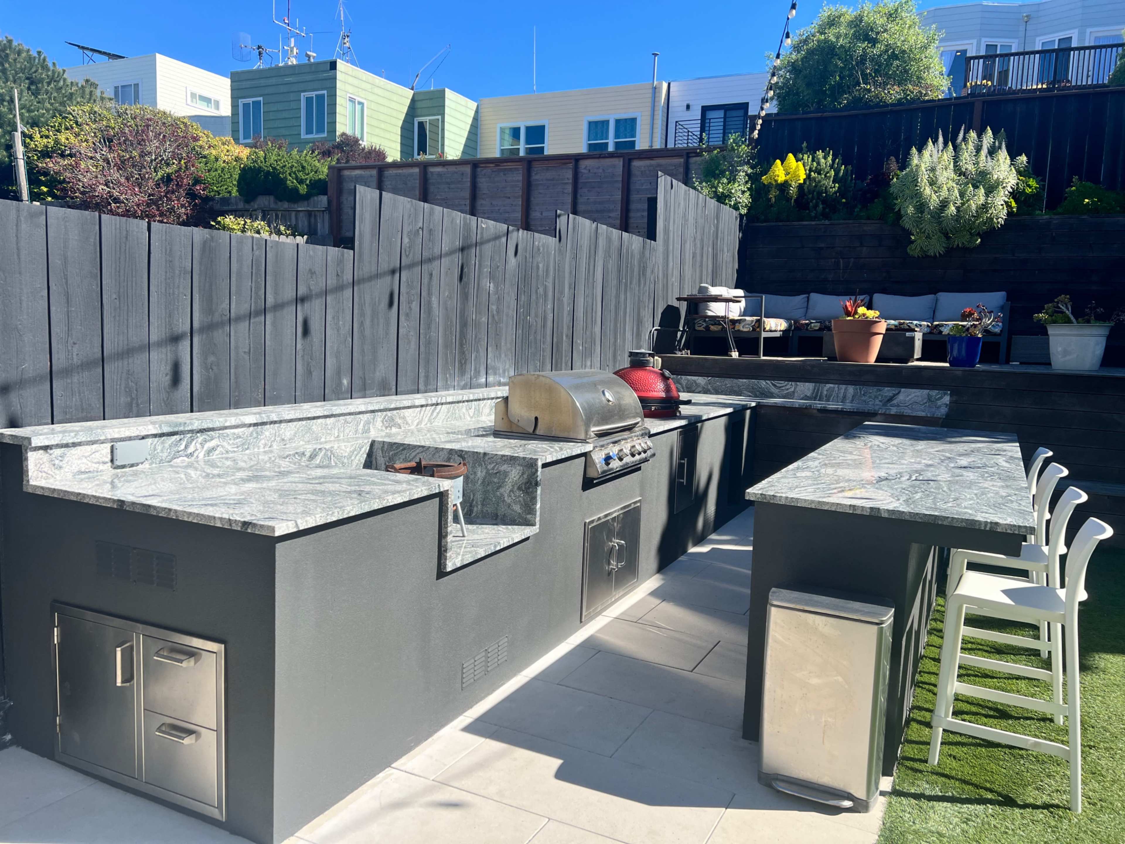 The image shows an outdoor kitchen equipped with a grill, stone countertops, and seating in a private backyard setting.