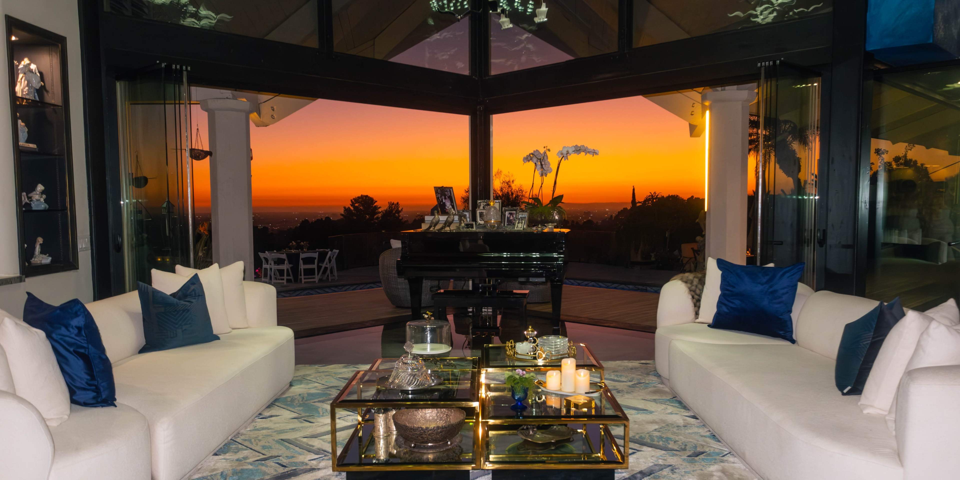 A modern living room features two white sofas facing a glass wall that showcases a vibrant sunset.