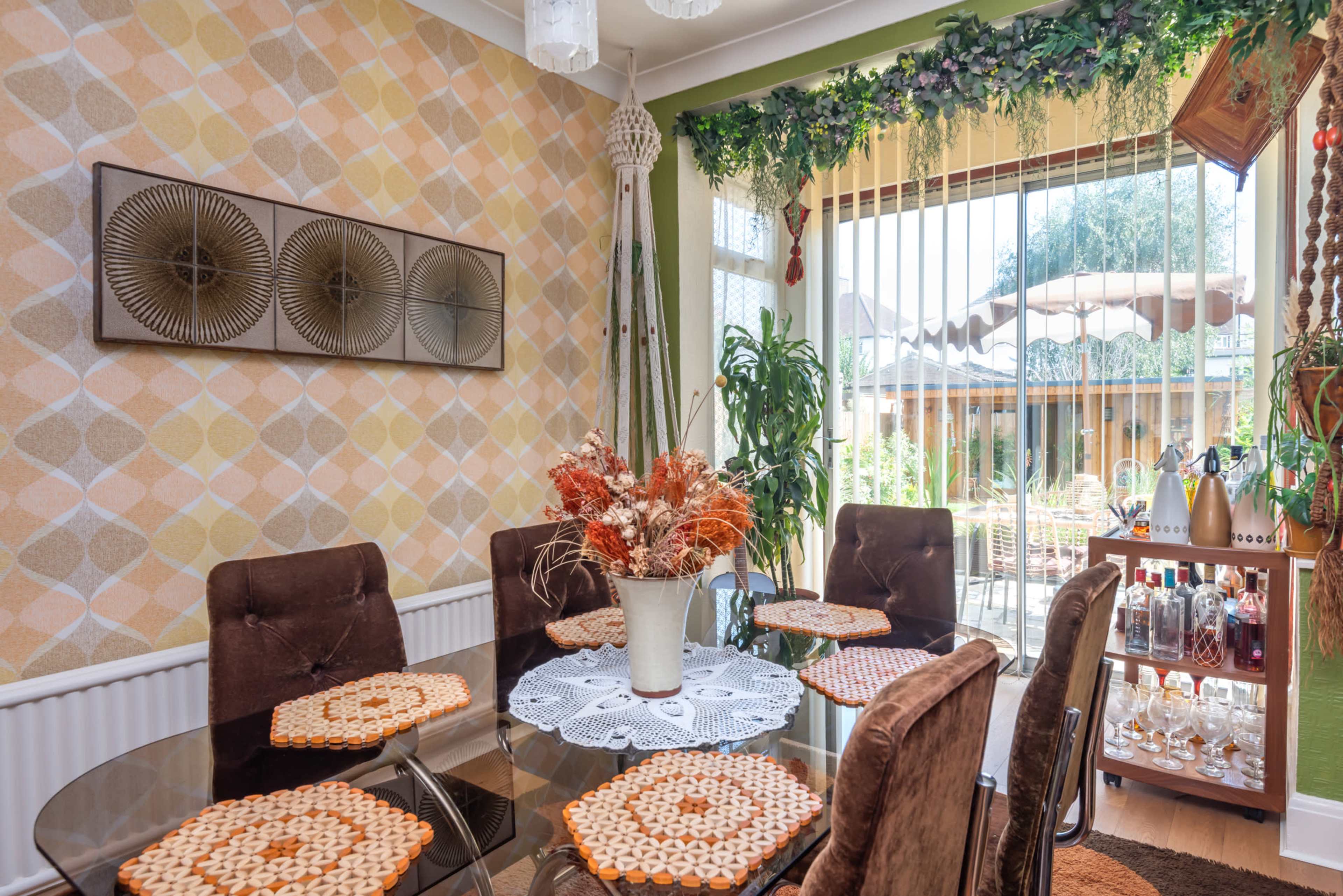 A dining room features a glass table surrounded by upholstered chairs, with decorative plants and patterned wallpaper in the background.
