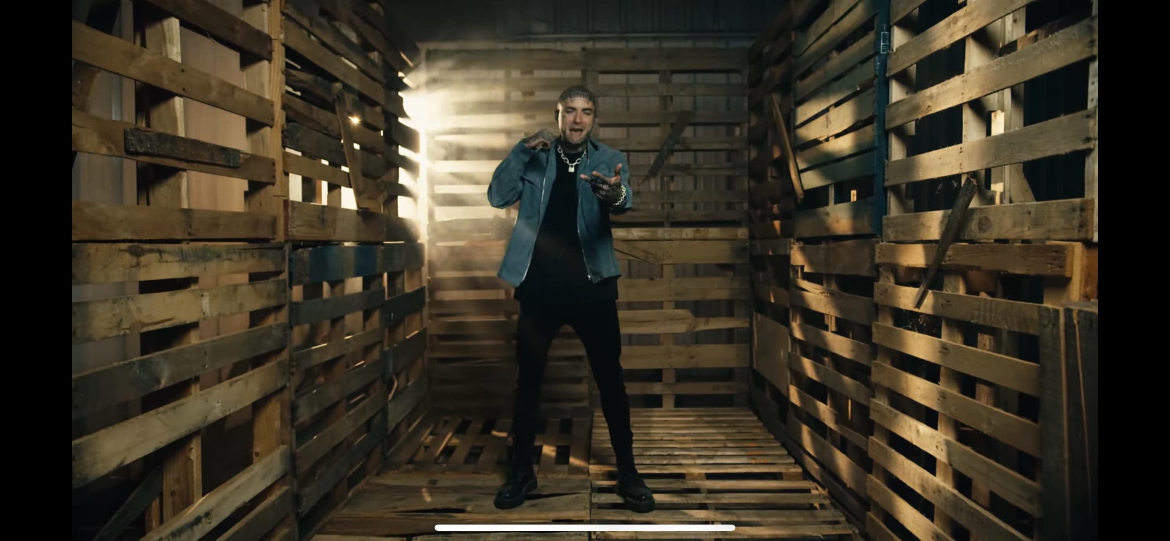 A person stands in a dimly lit wooden structure made of pallets, holding a microphone in a performance stance.