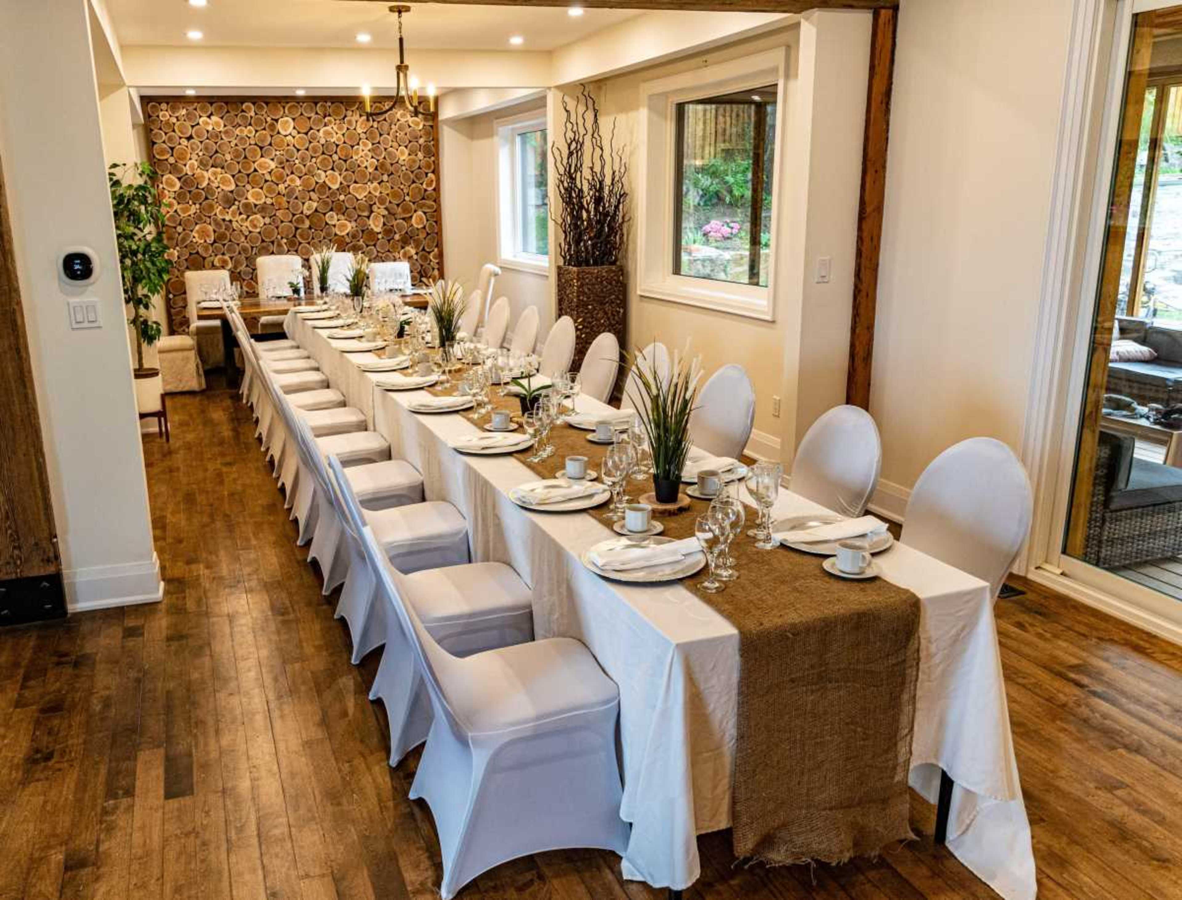 A long dining table is set with white tablecloths, plates, glasses, and decorations, arranged in a modern room with wooden flooring and a decorative wall.