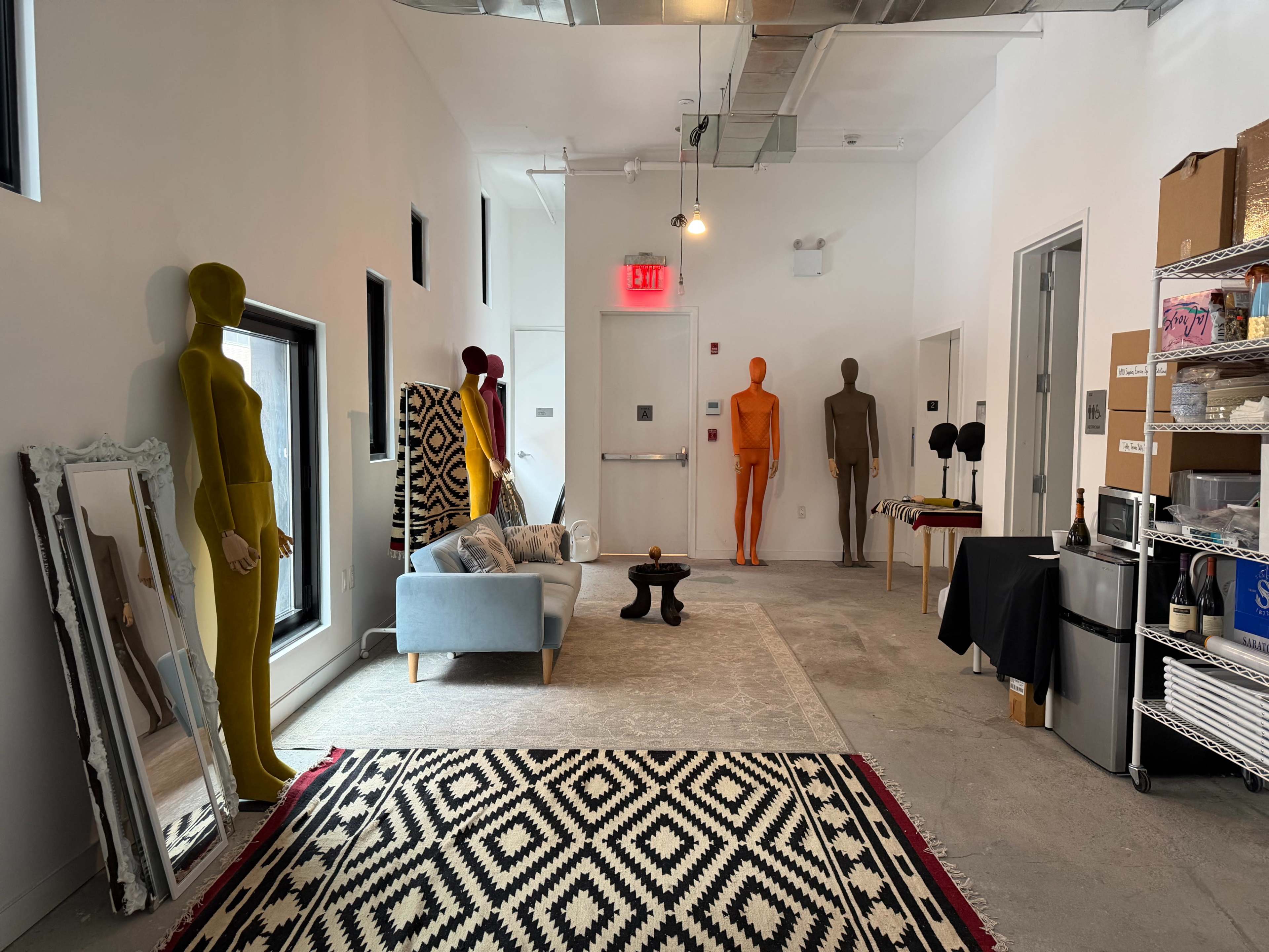 The scene shows a well-lit room with mannequins in various colors positioned around a seating area, featuring a rug, a small table, and art supplies.