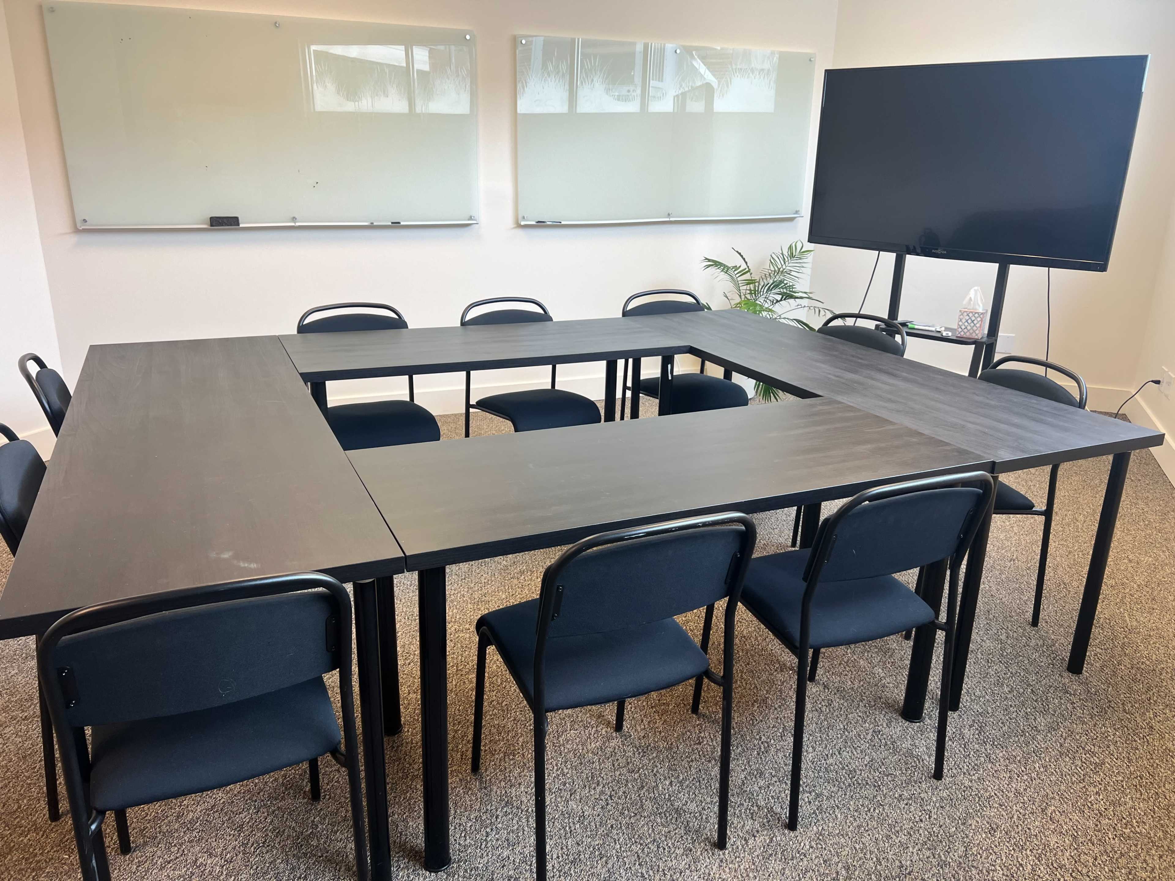 A conference room features a large, square table surrounded by eight chairs, with a television and whiteboards on the walls.