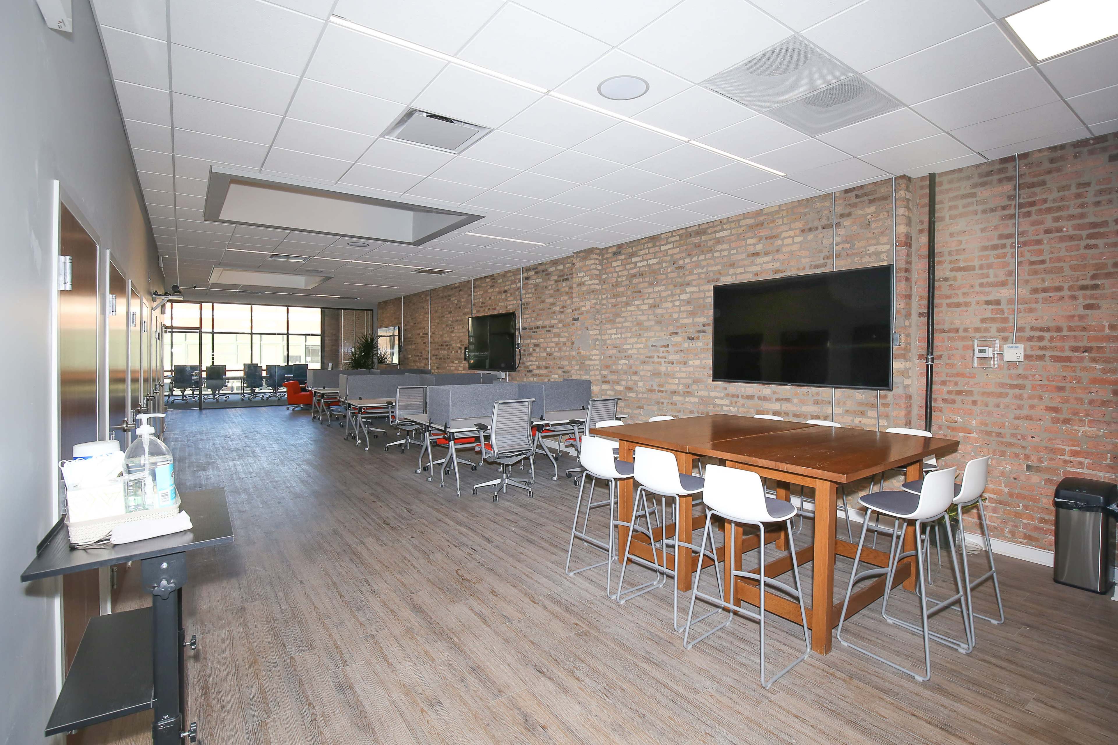 A modern office space with a long wooden table, chairs, and a lounge area featuring gray seating against exposed brick walls.