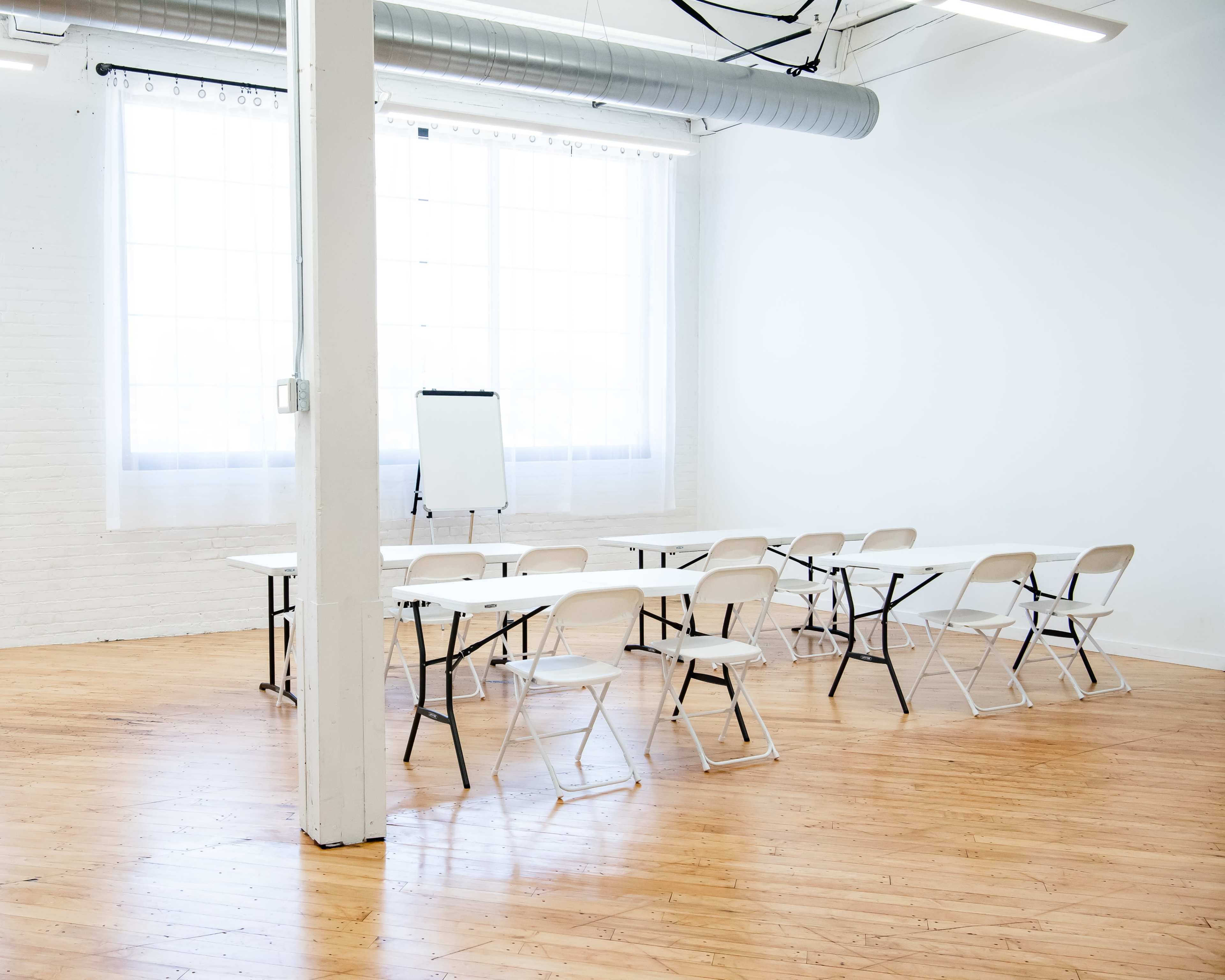 A spacious, brightly lit room features a row of folding chairs arranged around two long tables, with a whiteboard stand positioned at the front.