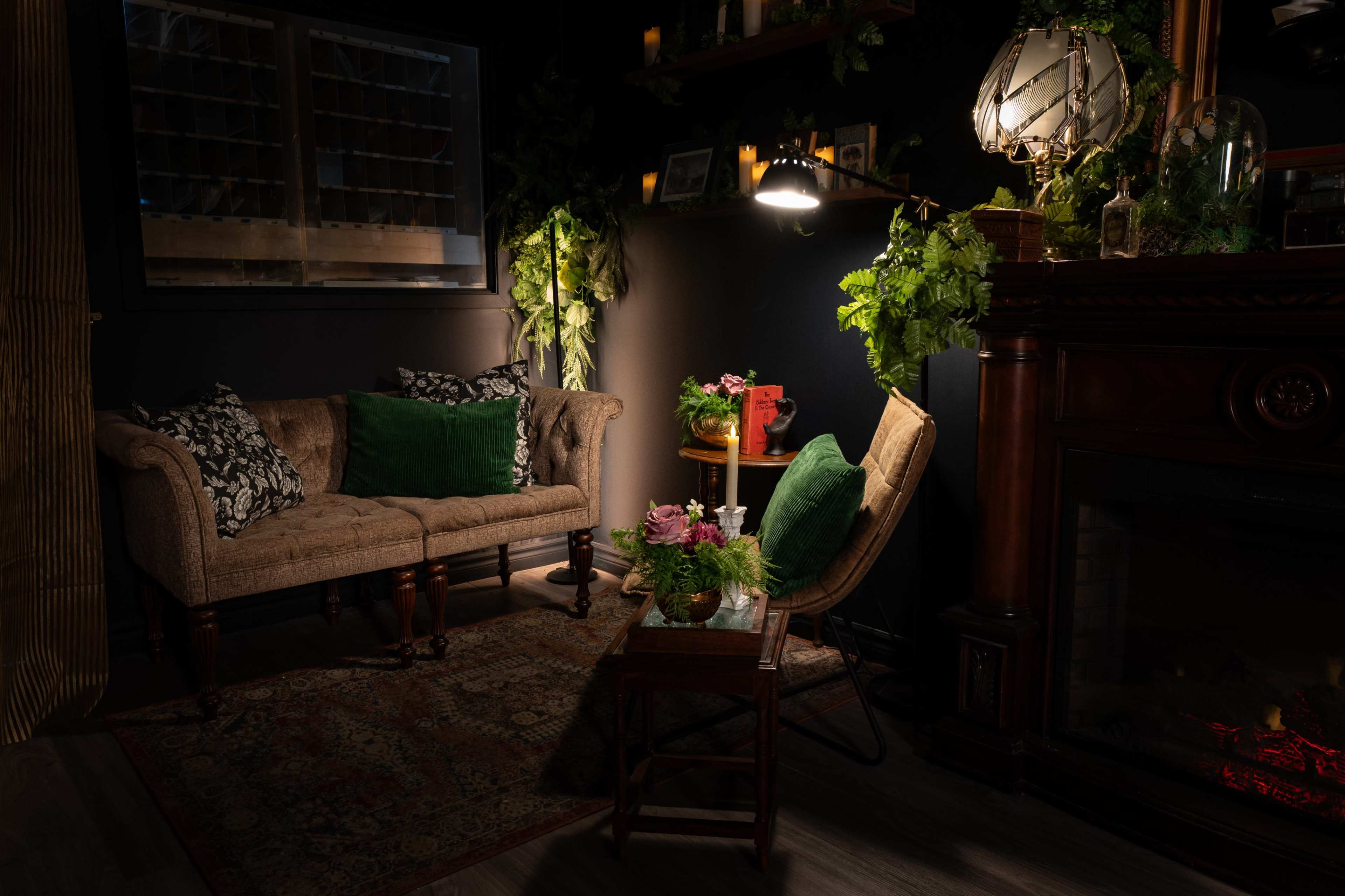 A cozy corner of a room features a vintage-style sofa, a small wooden table adorned with plants, and a lamp illuminating a dark space near a fireplace.