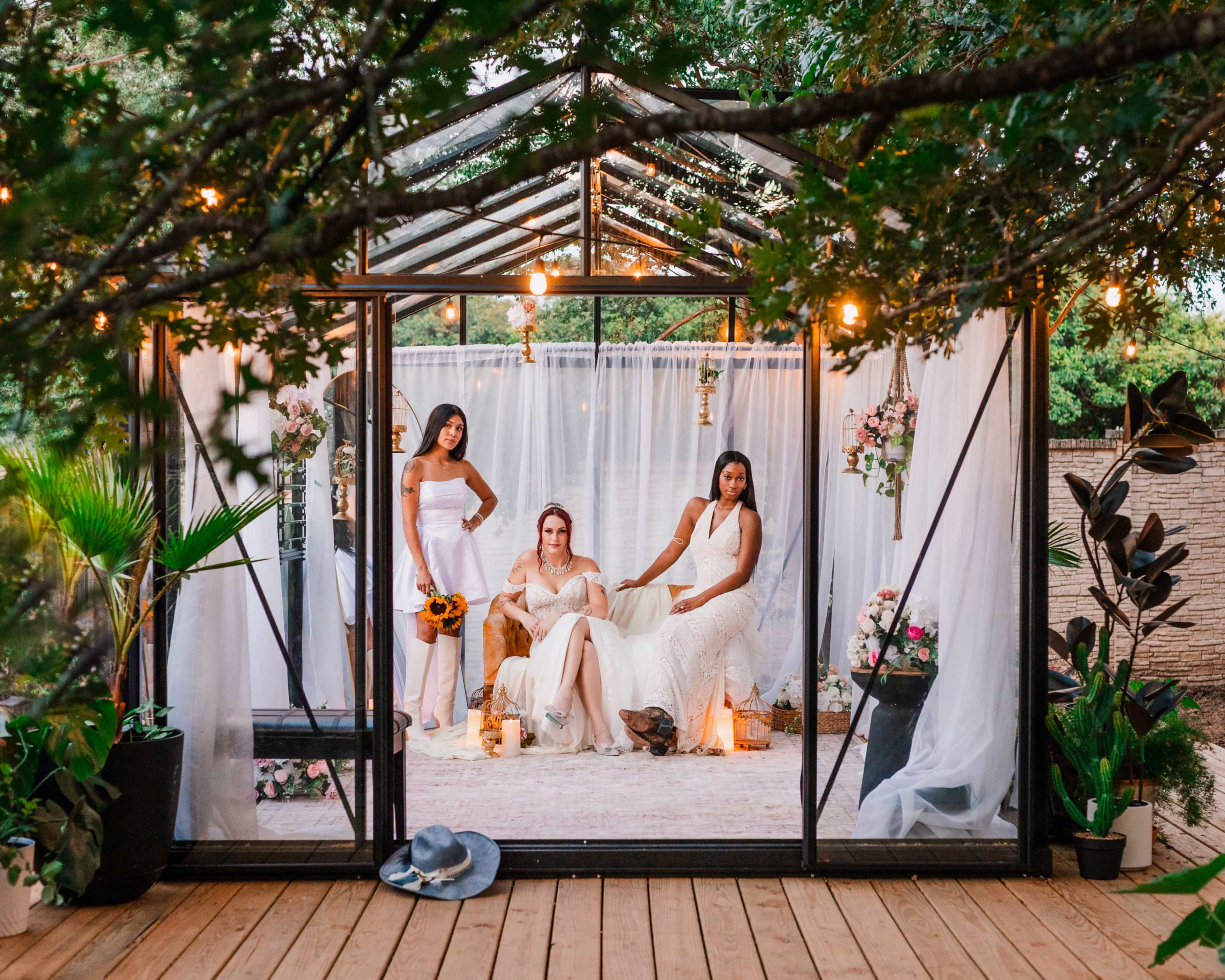 Three women dressed in white gowns pose inside a glass greenhouse adorned with plants and decorative accents.