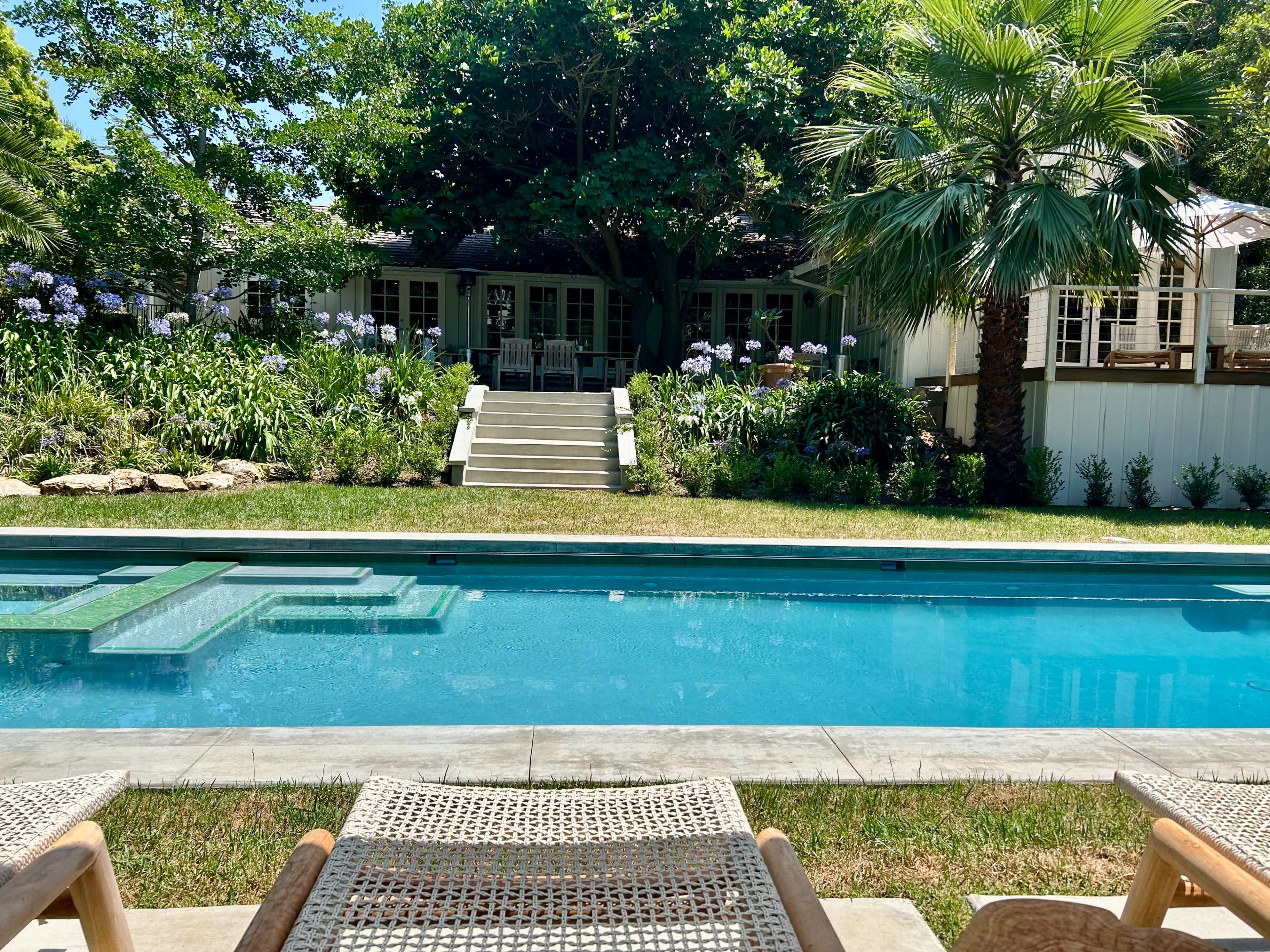 A clear pool reflects the surrounding greenery and a house in the background, framed by lounge chairs at the water's edge.