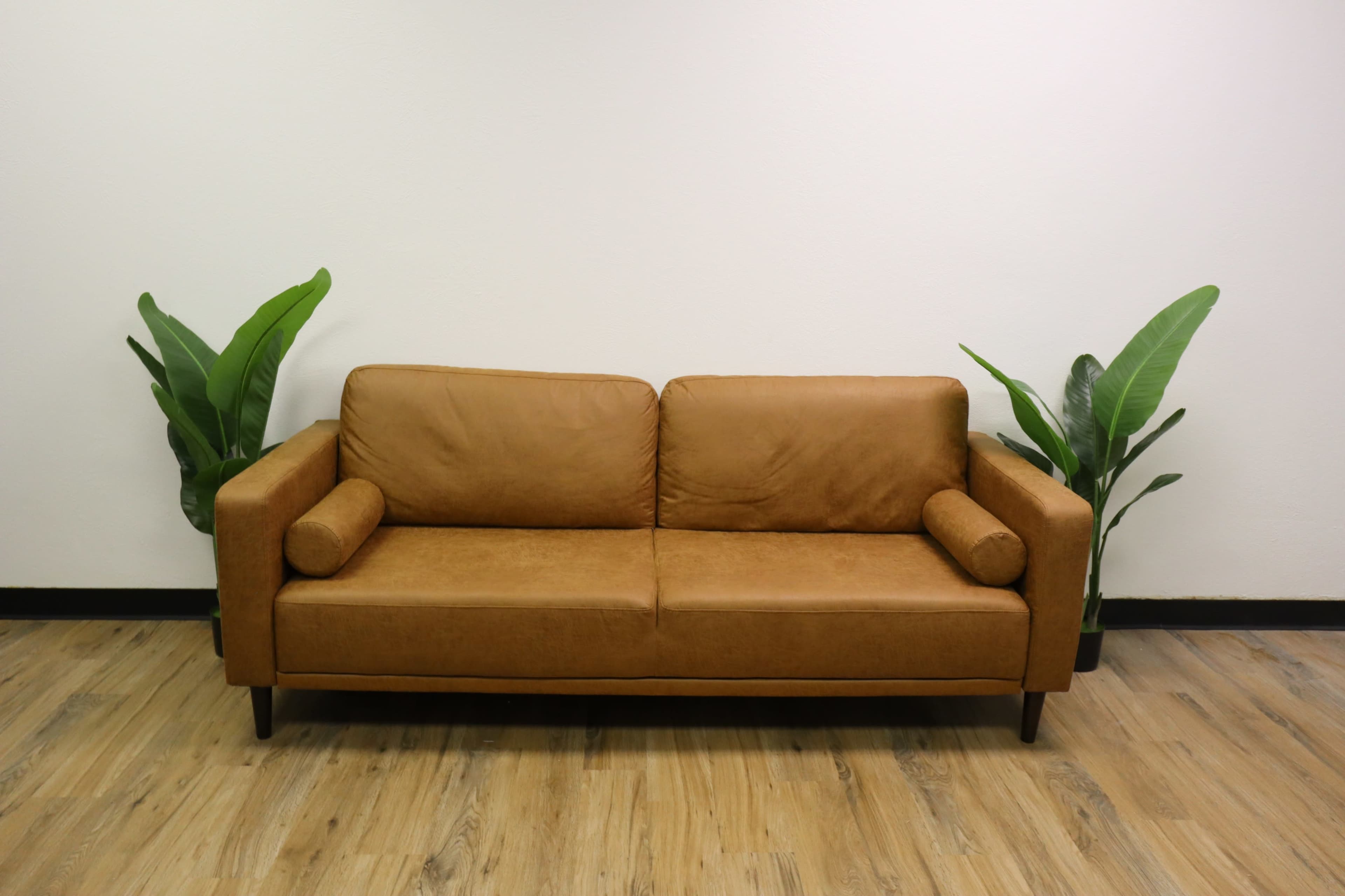 A brown leather sofa is centered against a white wall, flanked by two green potted plants.