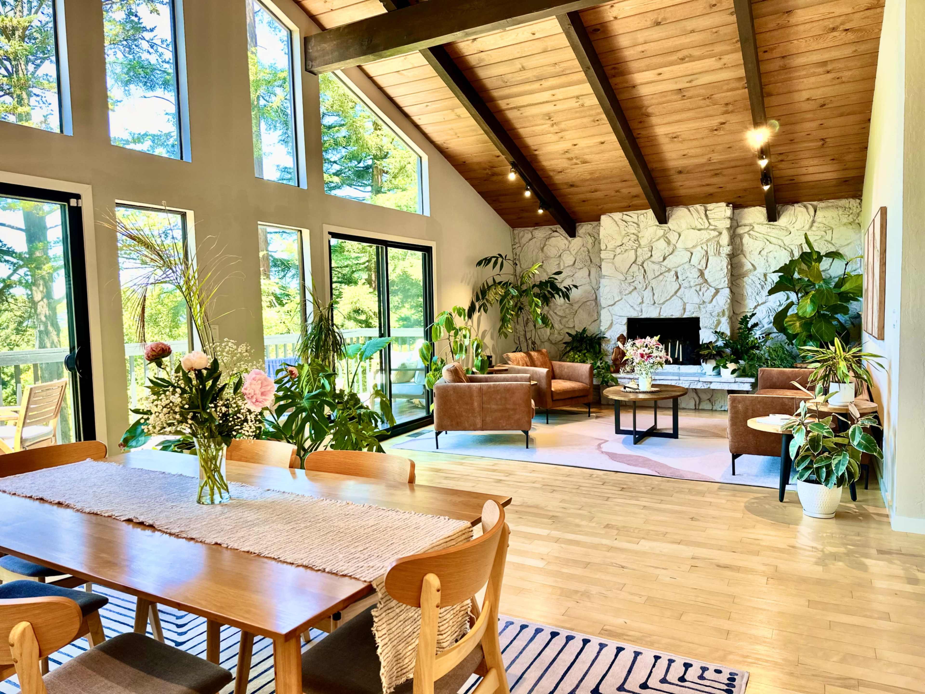 A spacious living area features large windows, wooden beams, and a blend of modern and natural decor, including a dining table and comfortable seating surrounded by greenery.