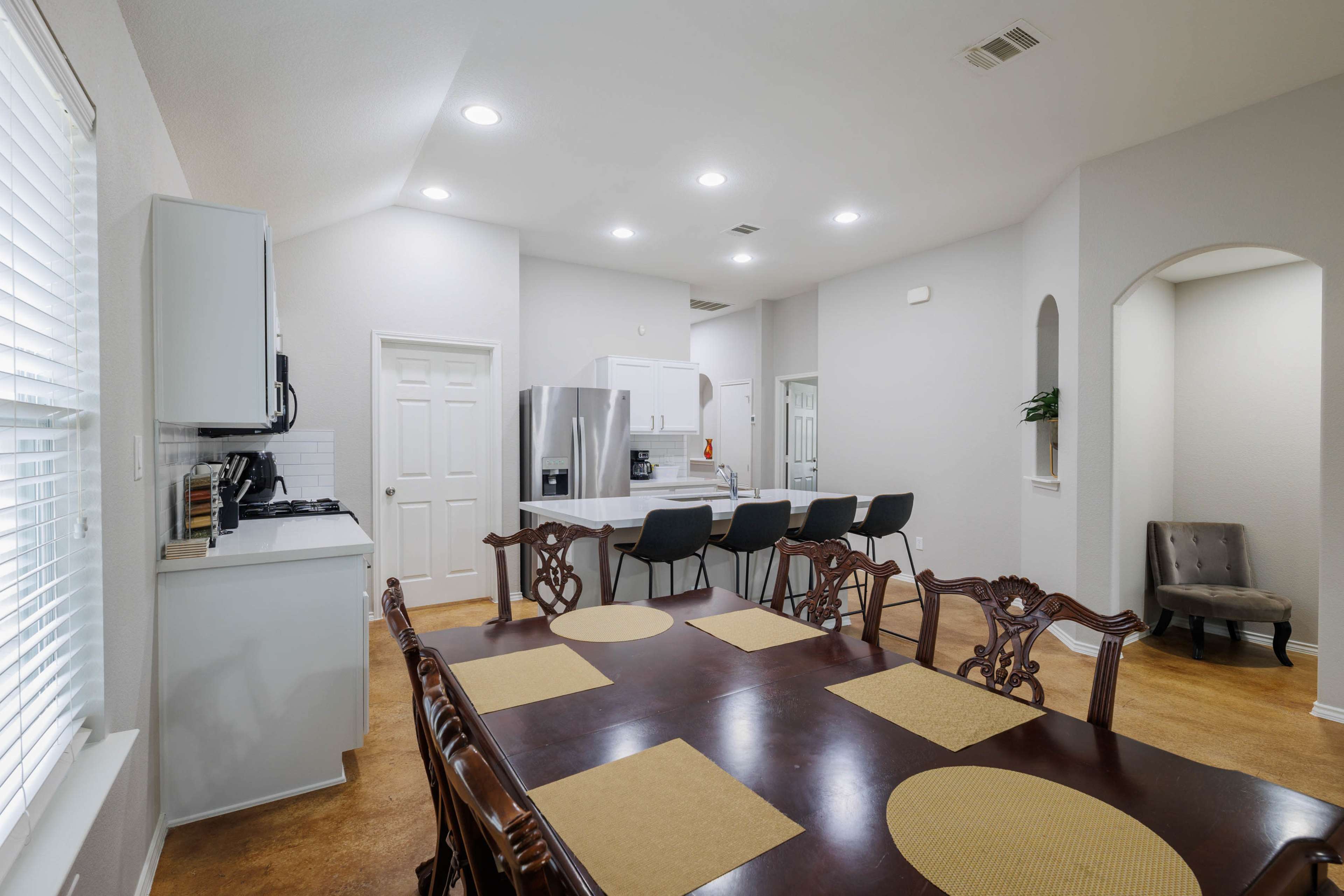 A well-lit kitchen and dining area features a wooden table with placemats, bar seating at the kitchen counter, and modern appliances.