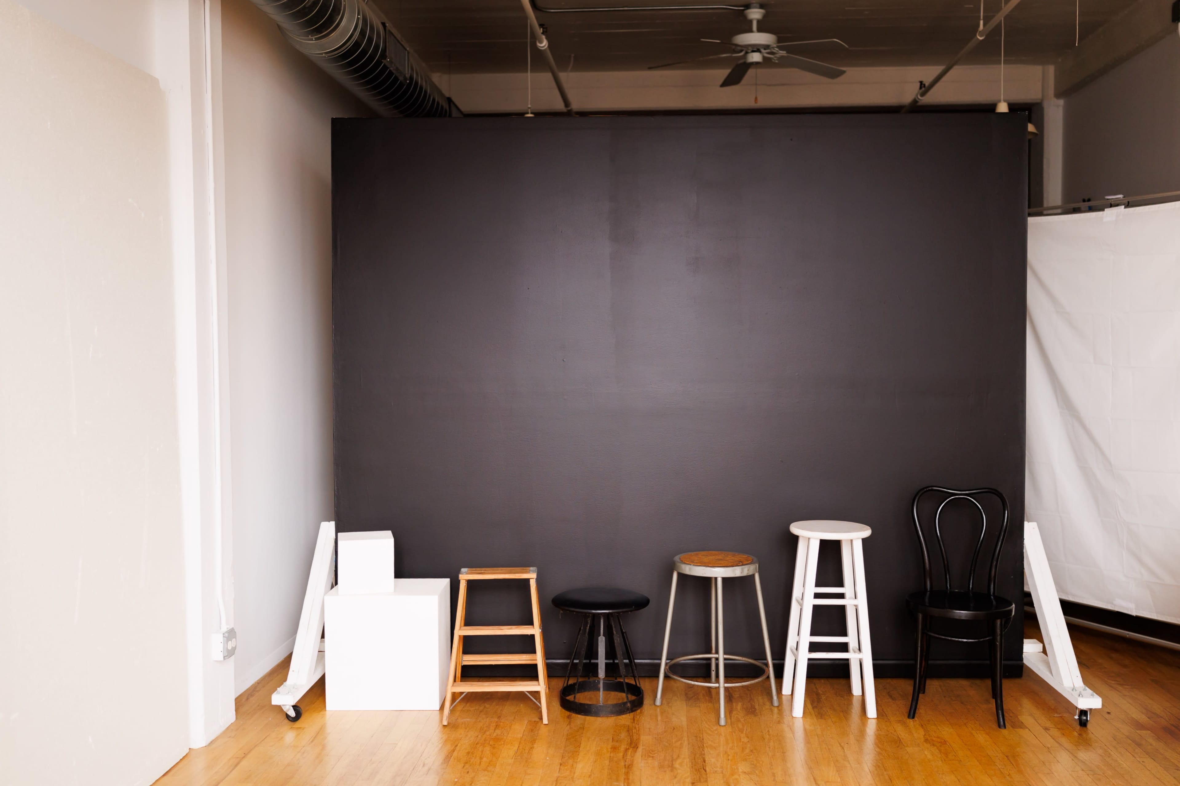 The image shows a minimalistic studio space with a dark gray backdrop and five different stools lined up against it.