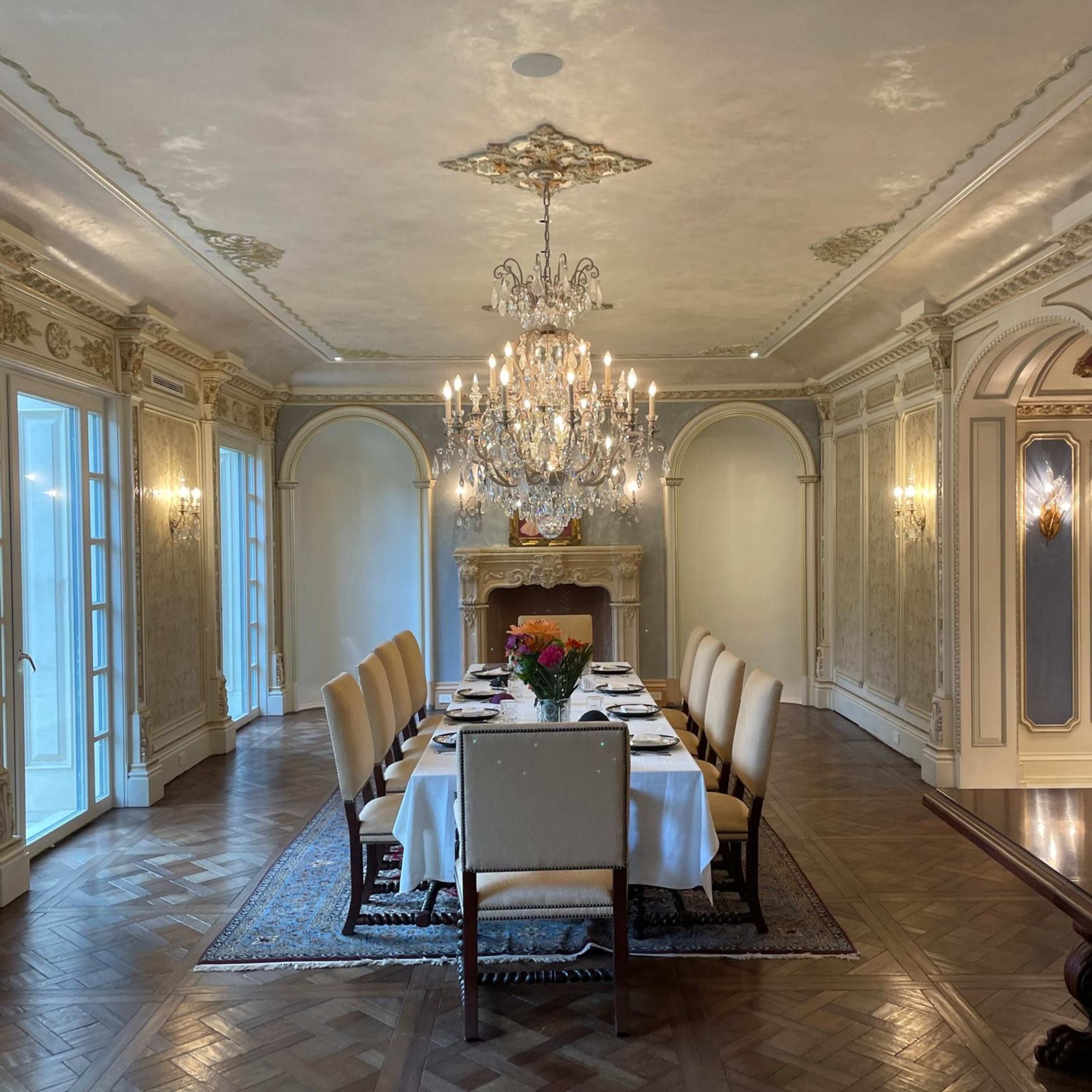 An elegantly decorated dining room with a long table set for a meal, surrounded by ten upholstered chairs and adorned with a large chandelier above.