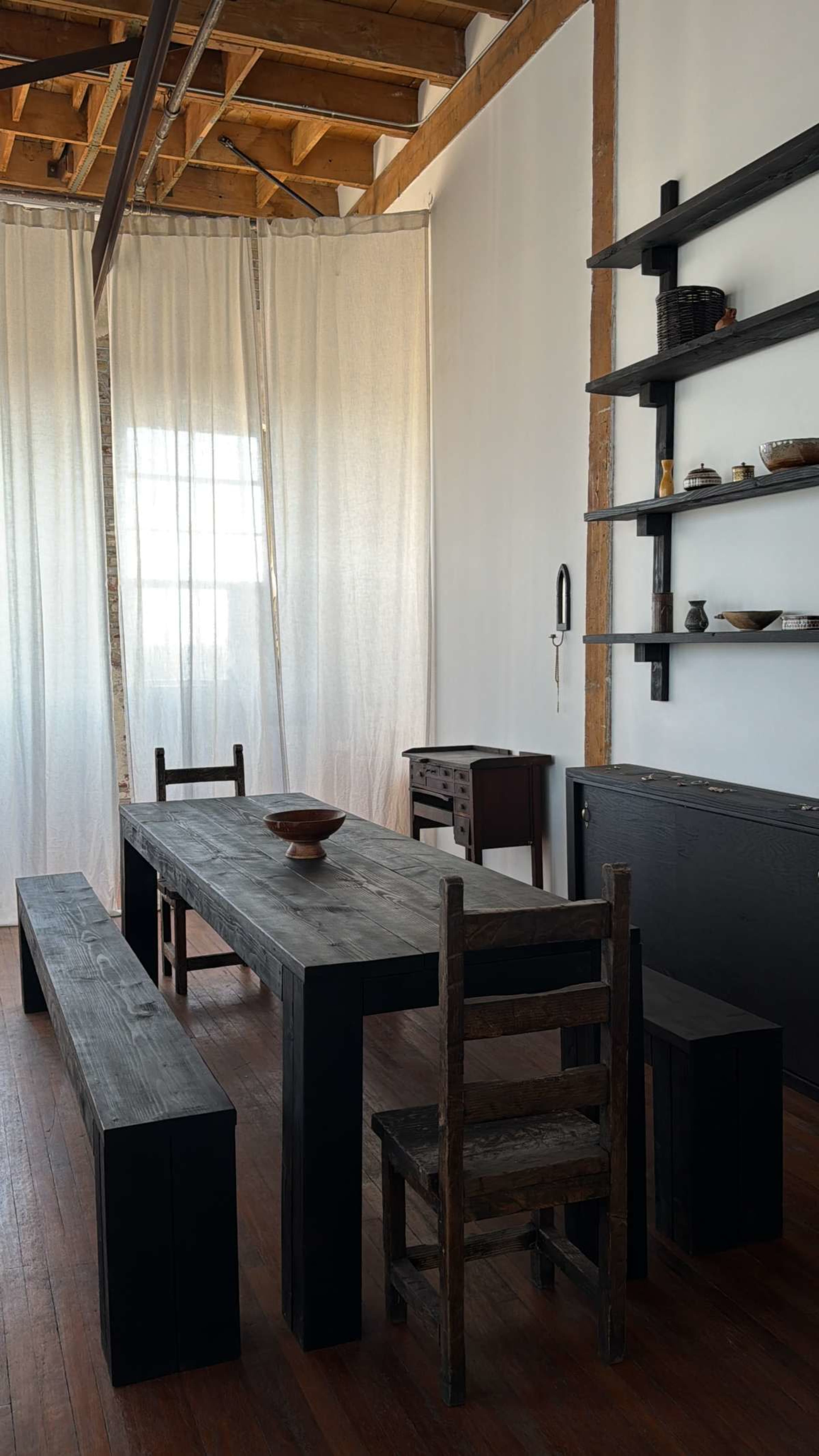 The image shows a minimalist dining area featuring a large black wooden table, a couple of benches, and a small chest of drawers, all set against a backdrop of white curtains and exposed wooden beams.