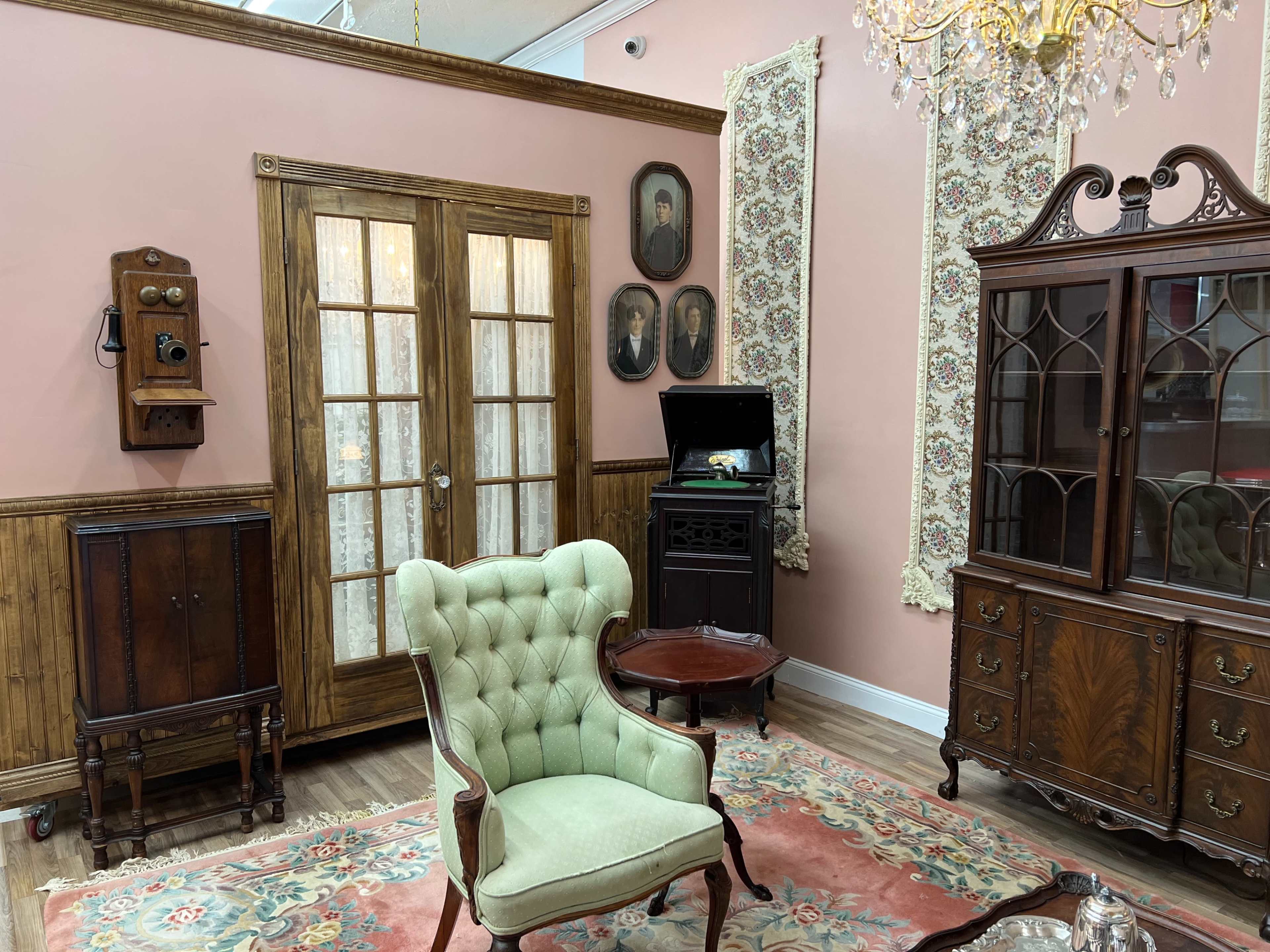 The scene shows a vintage room with a green upholstered armchair, a wooden table, a dark china cabinet, and an antique phone, all against a backdrop of pink walls and decorative elements.