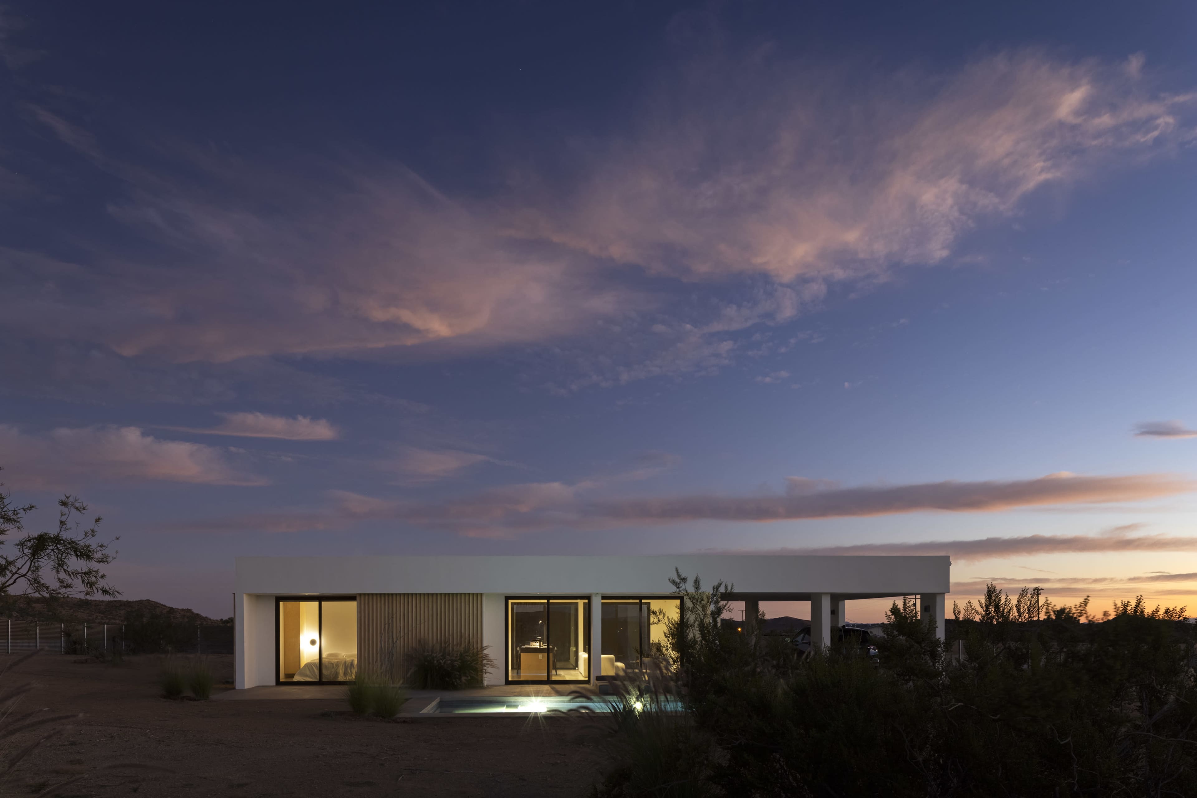 A modern one-story house with large windows is set against a twilight sky featuring clouds and sparse desert vegetation.