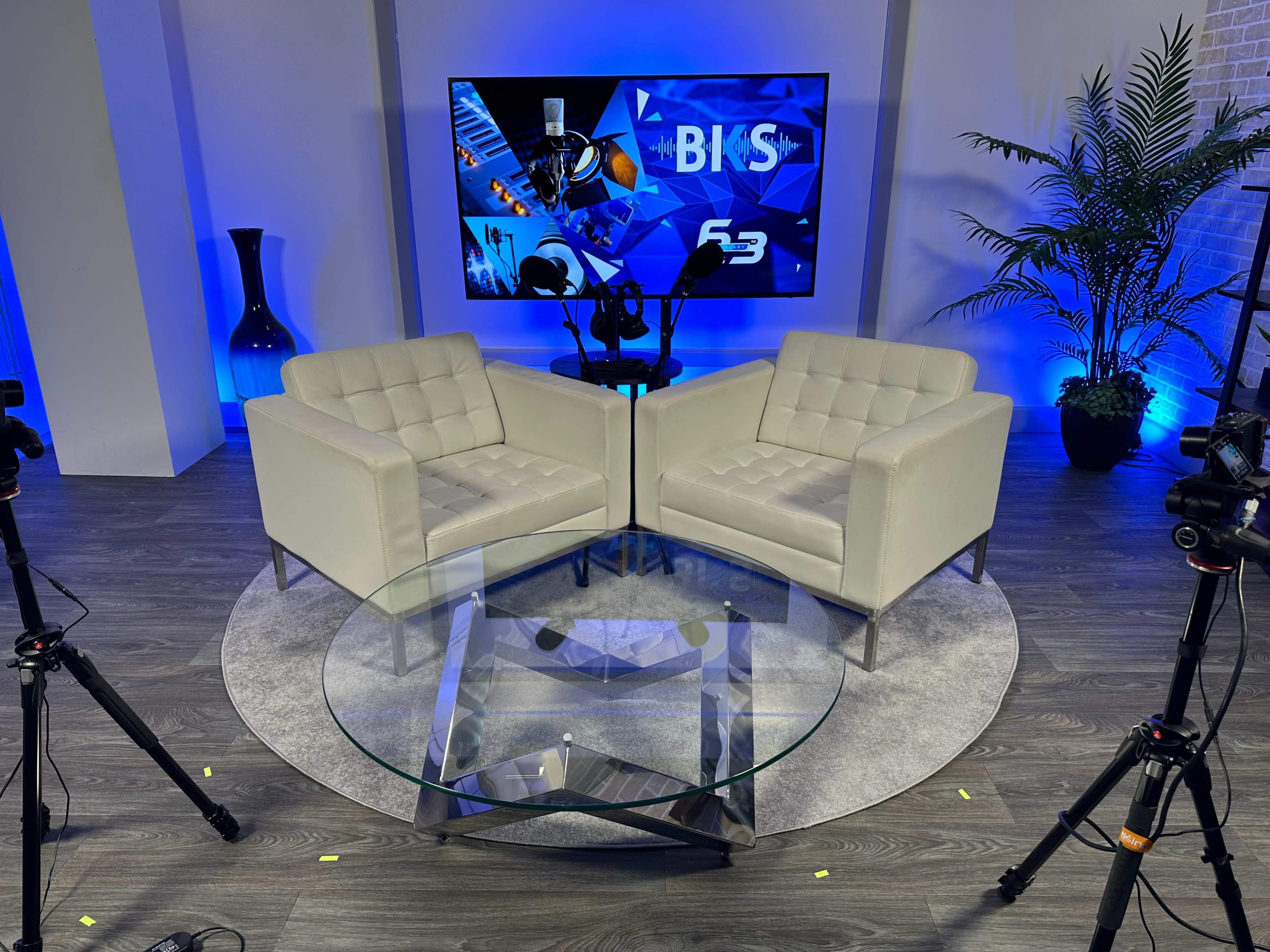 A modern interview setup with two white armchairs facing each other around a glass coffee table, framed by a blue-lit backdrop featuring a screen displaying graphics.