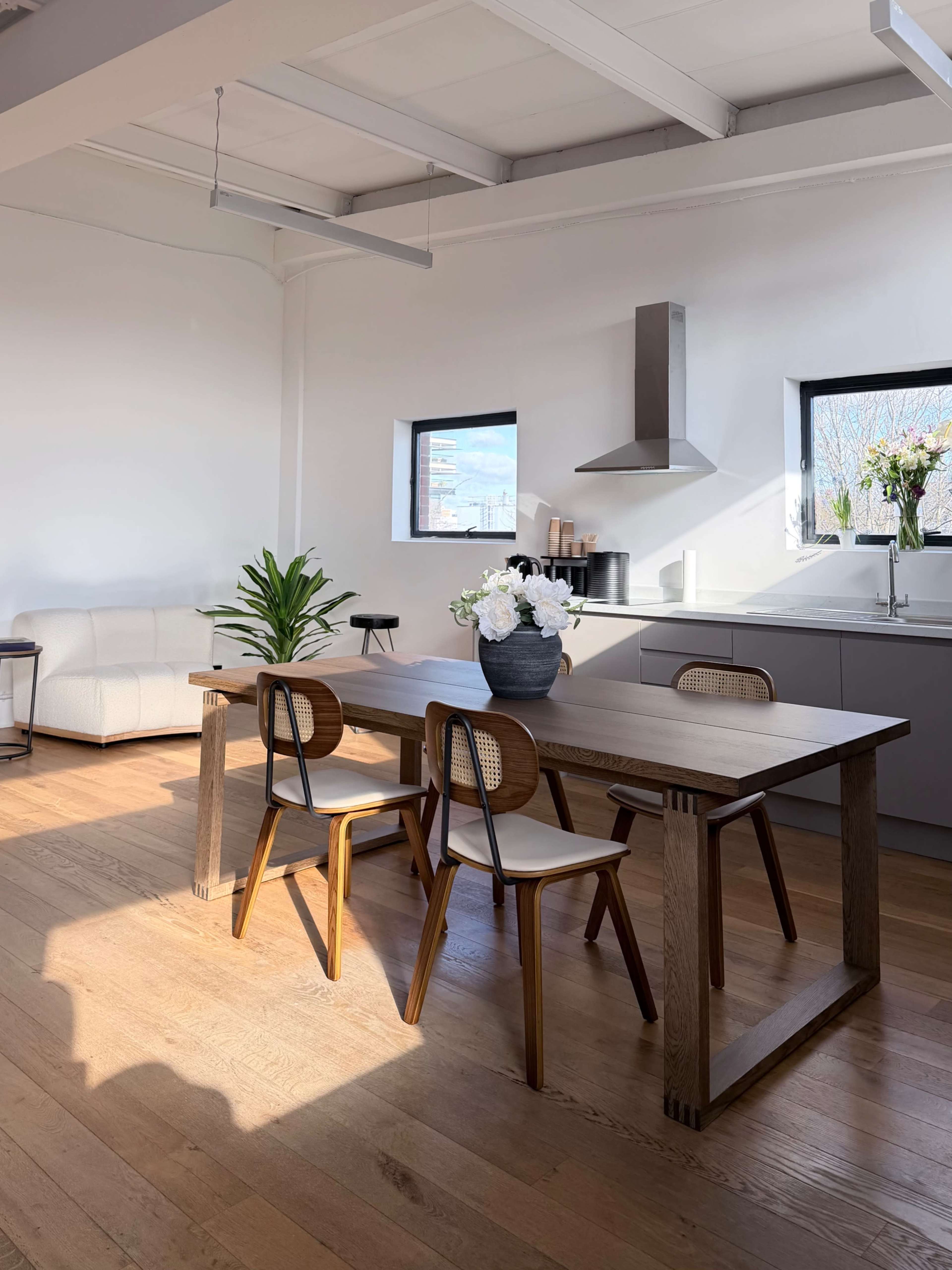 A modern kitchen and dining area features a wooden table with three chairs, a potted plant, and a vase of flowers, illuminated by natural light from windows.