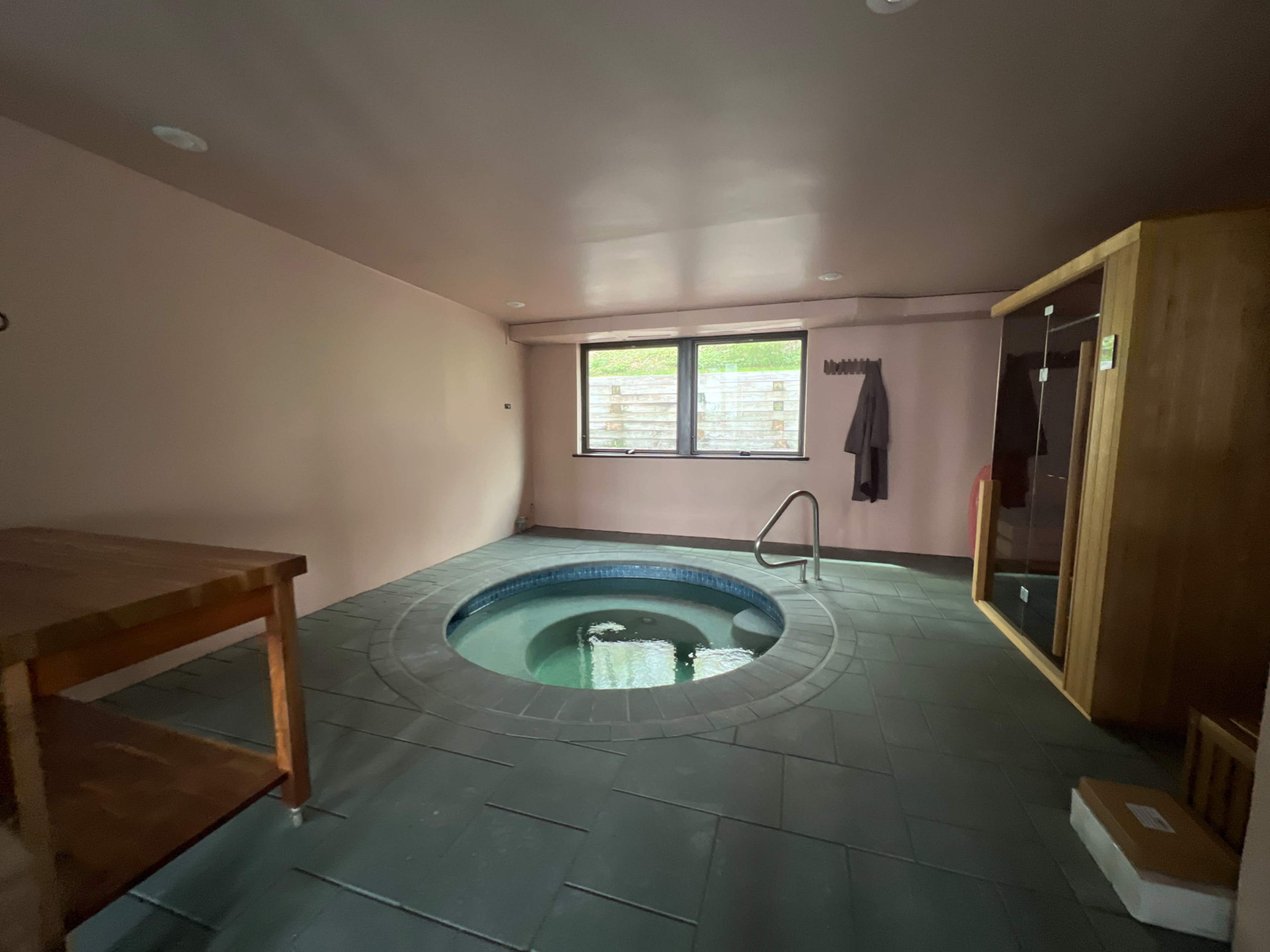 The image shows a spacious indoor spa area featuring a circular hot tub, a wooden bench, and a glass-enclosed shower.