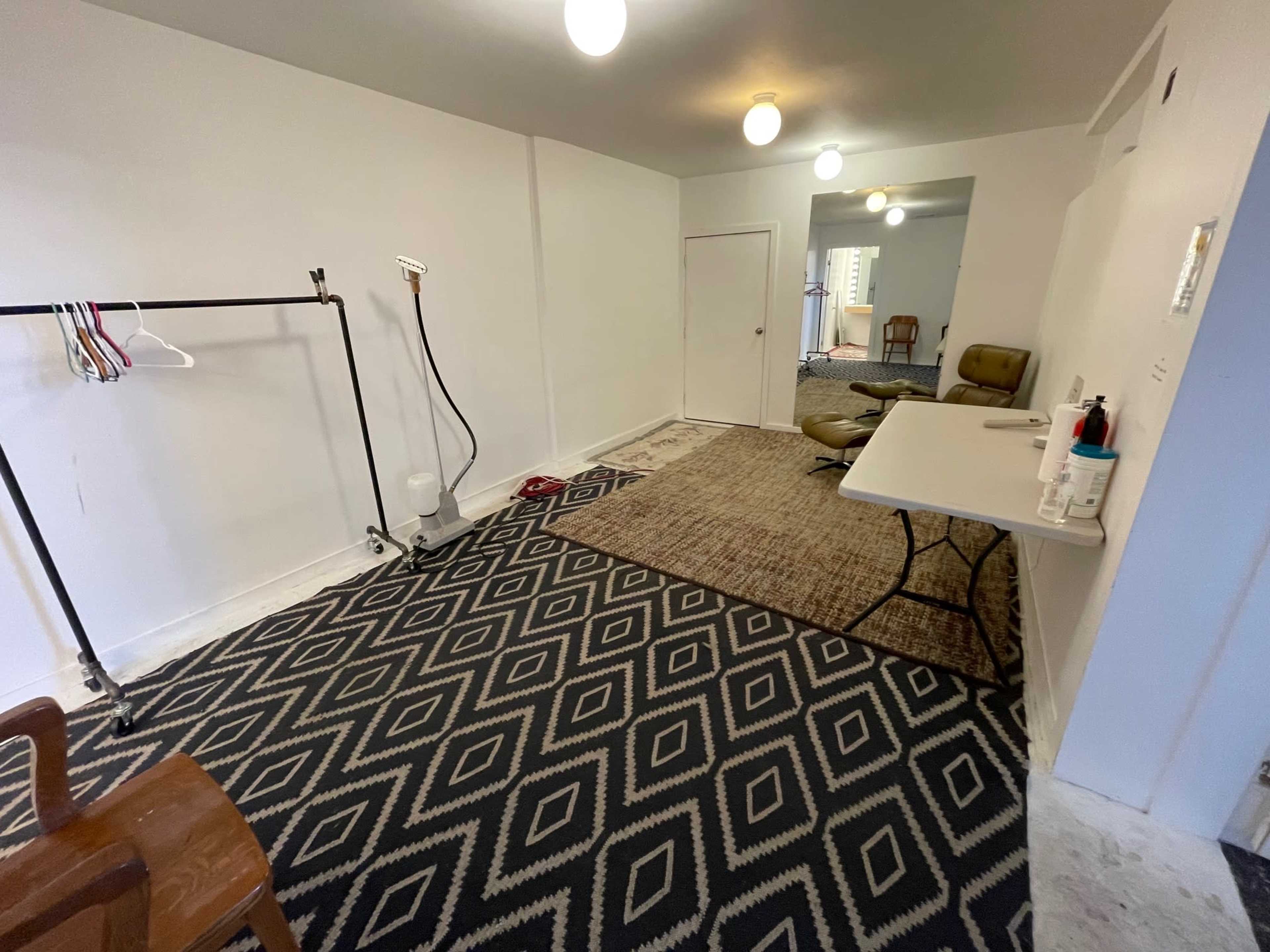 The image shows an empty room with patterned carpeting, a clothing rack, a table, and two light fixtures hanging from the ceiling.
